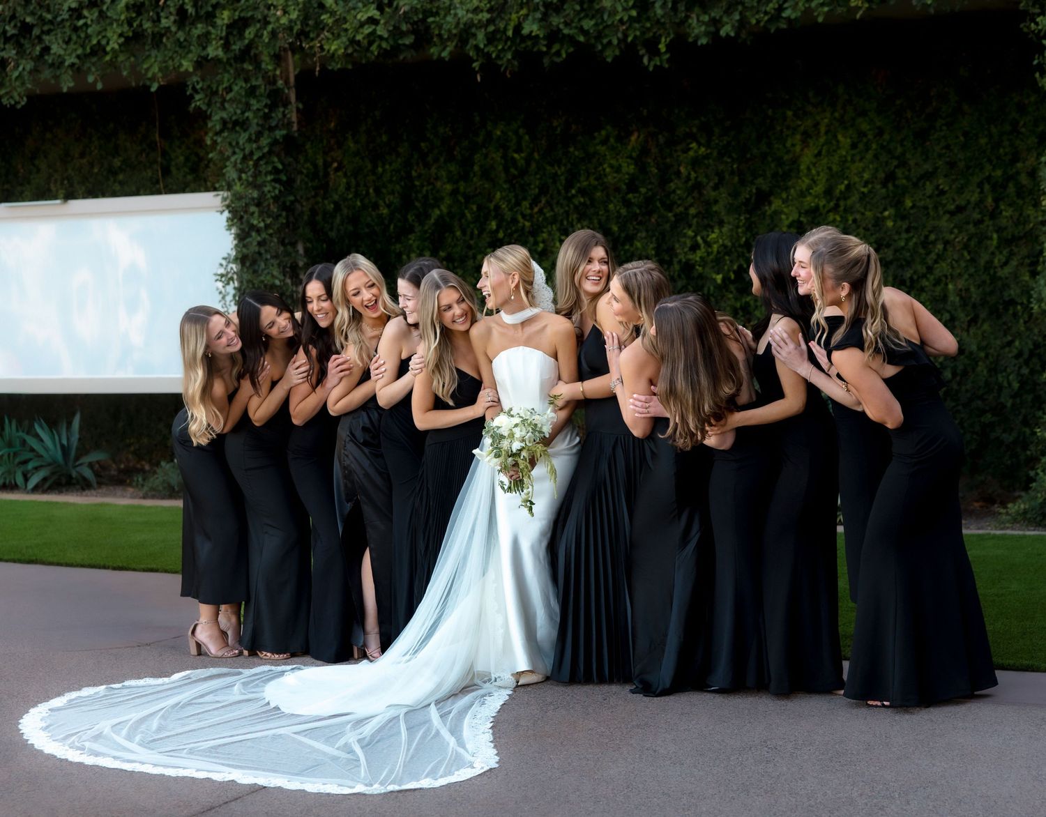 Bride in white gown with cathedral veil poses with bridesmaids in black dresses outdoors.