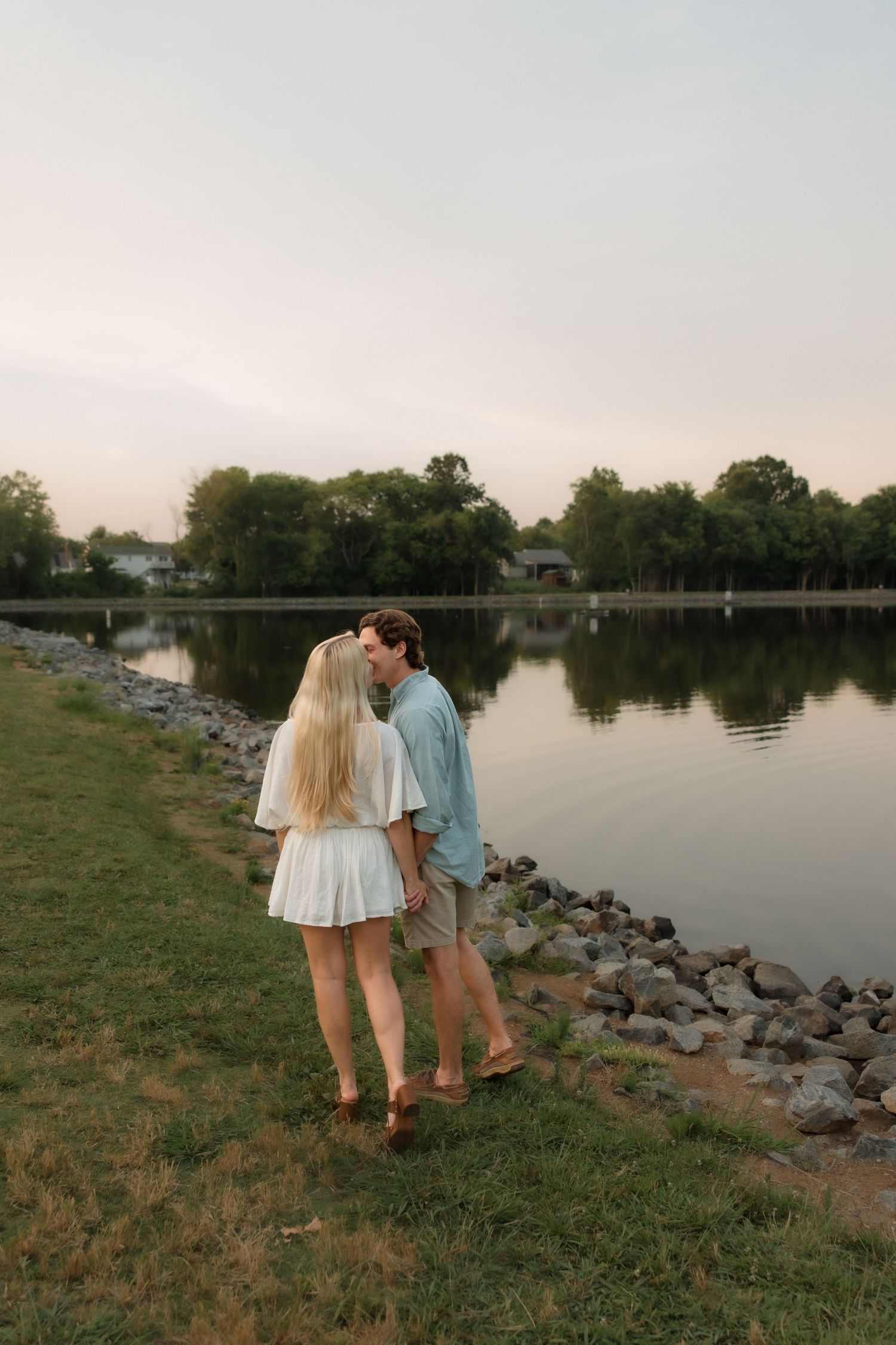 Couple sharing a kiss while standing on a grassy lakeside shore at dusk with calm water reflection.