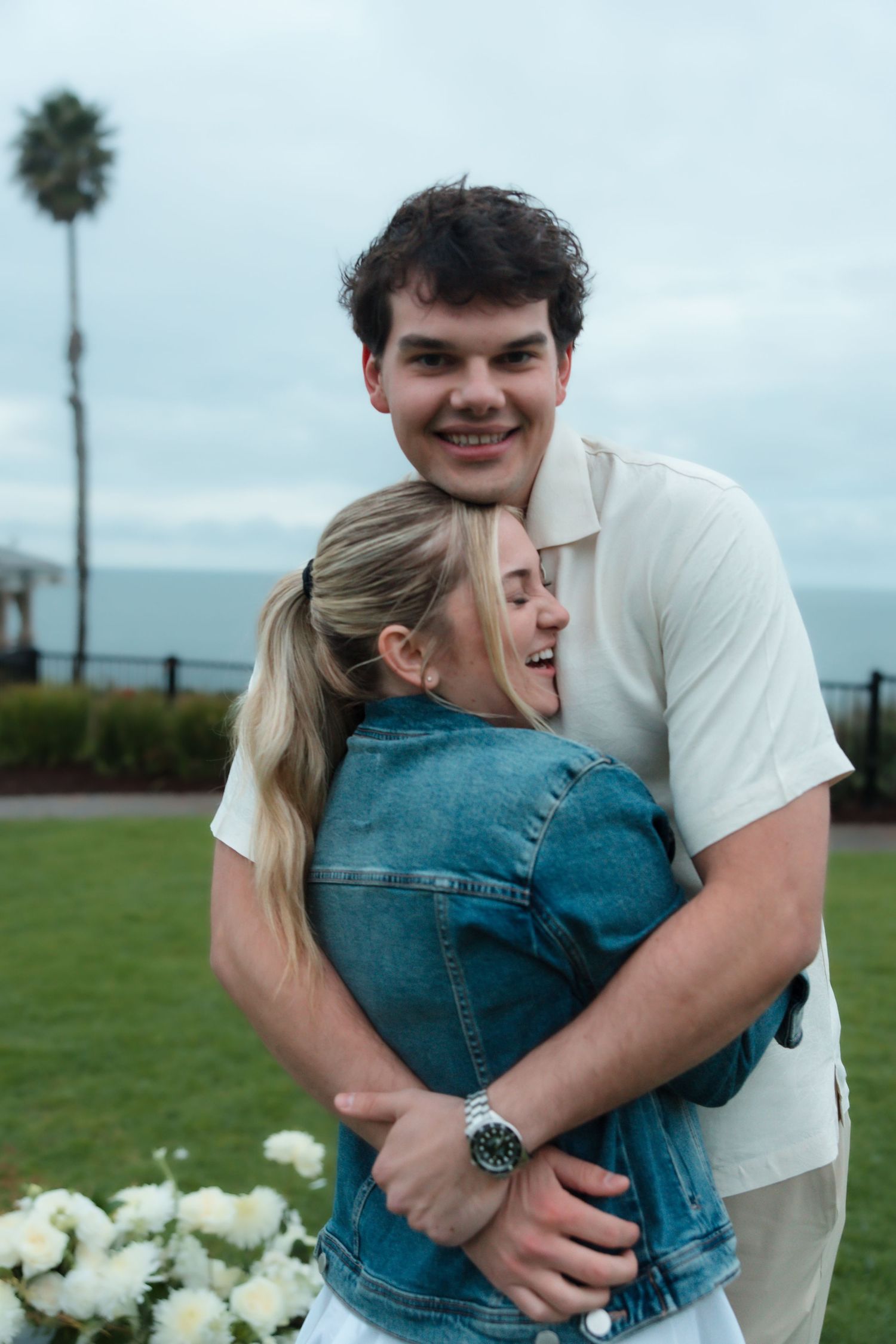 Smiling man hugging blonde woman in denim jacket near white flowers with ocean backdrop.