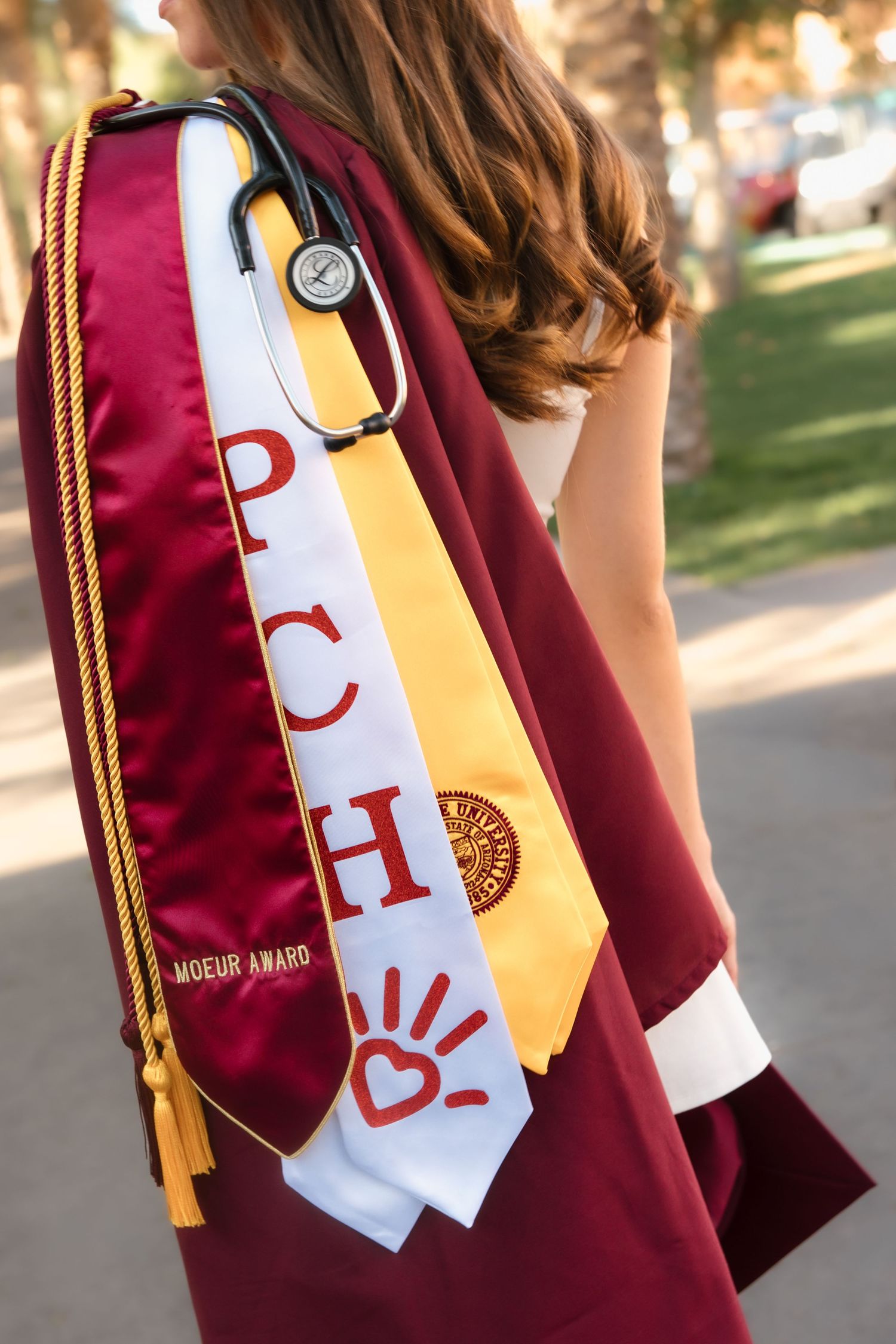 Graduate displays PCHO stole, Moeur Award sash, gold honor cords, and stethoscope on maroon gown.