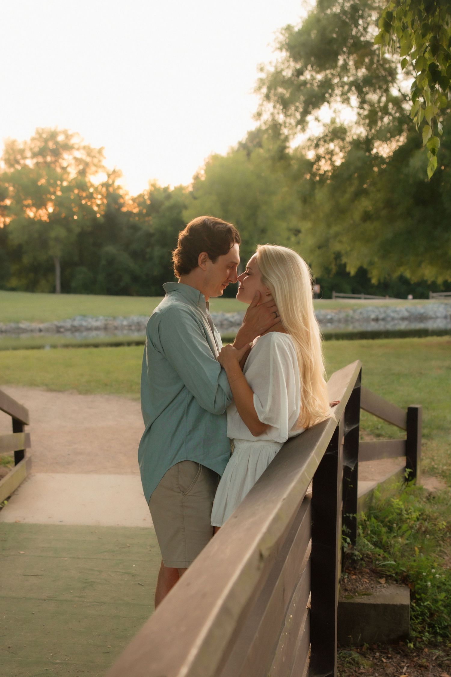 Couple sharing a romantic nose-to-nose moment on a wooden bridge surrounded by lush green trees at sunset.