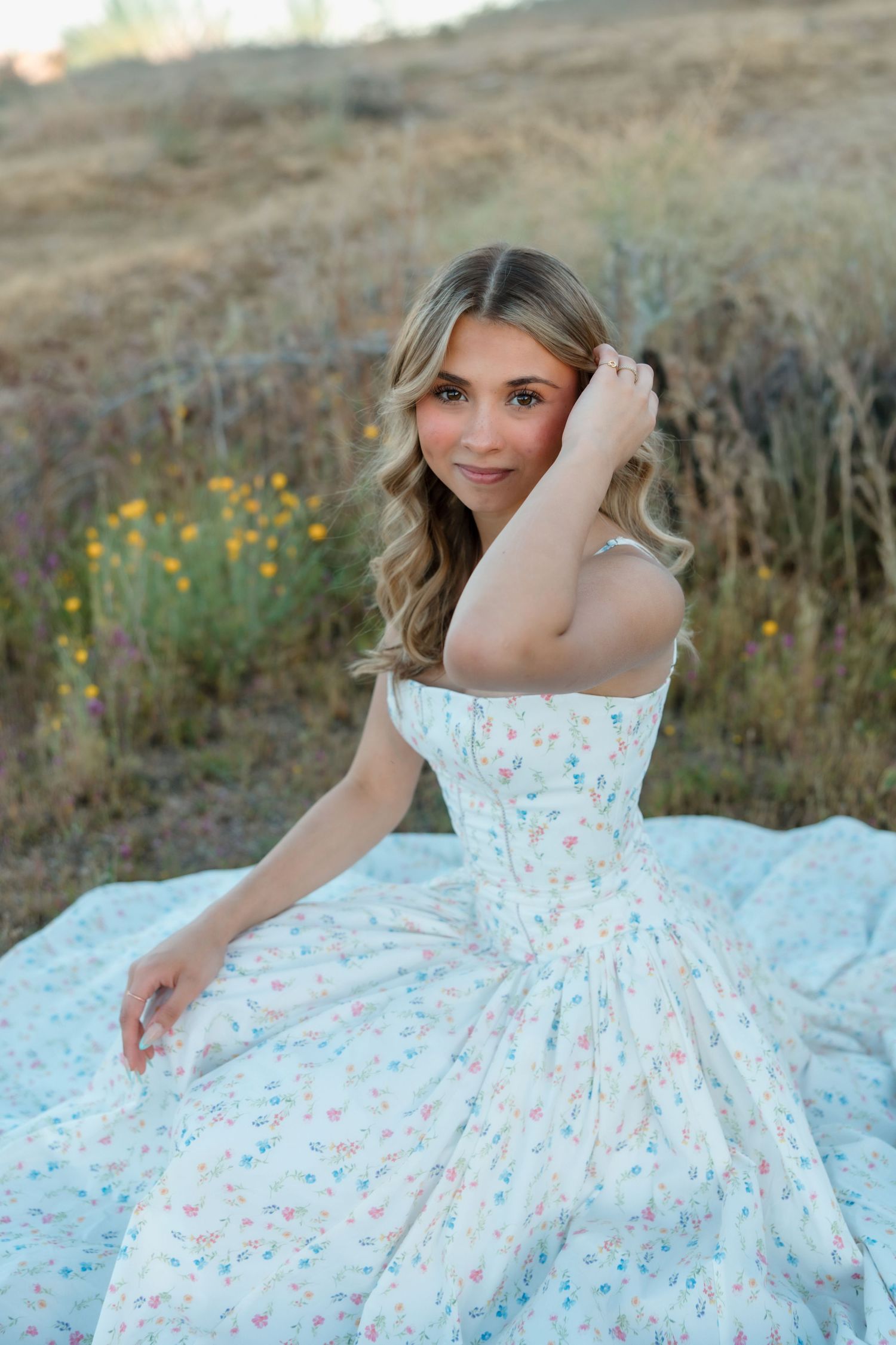 Young woman in floral sundress sitting outdoors in a field with wildflowers, posing gracefully.