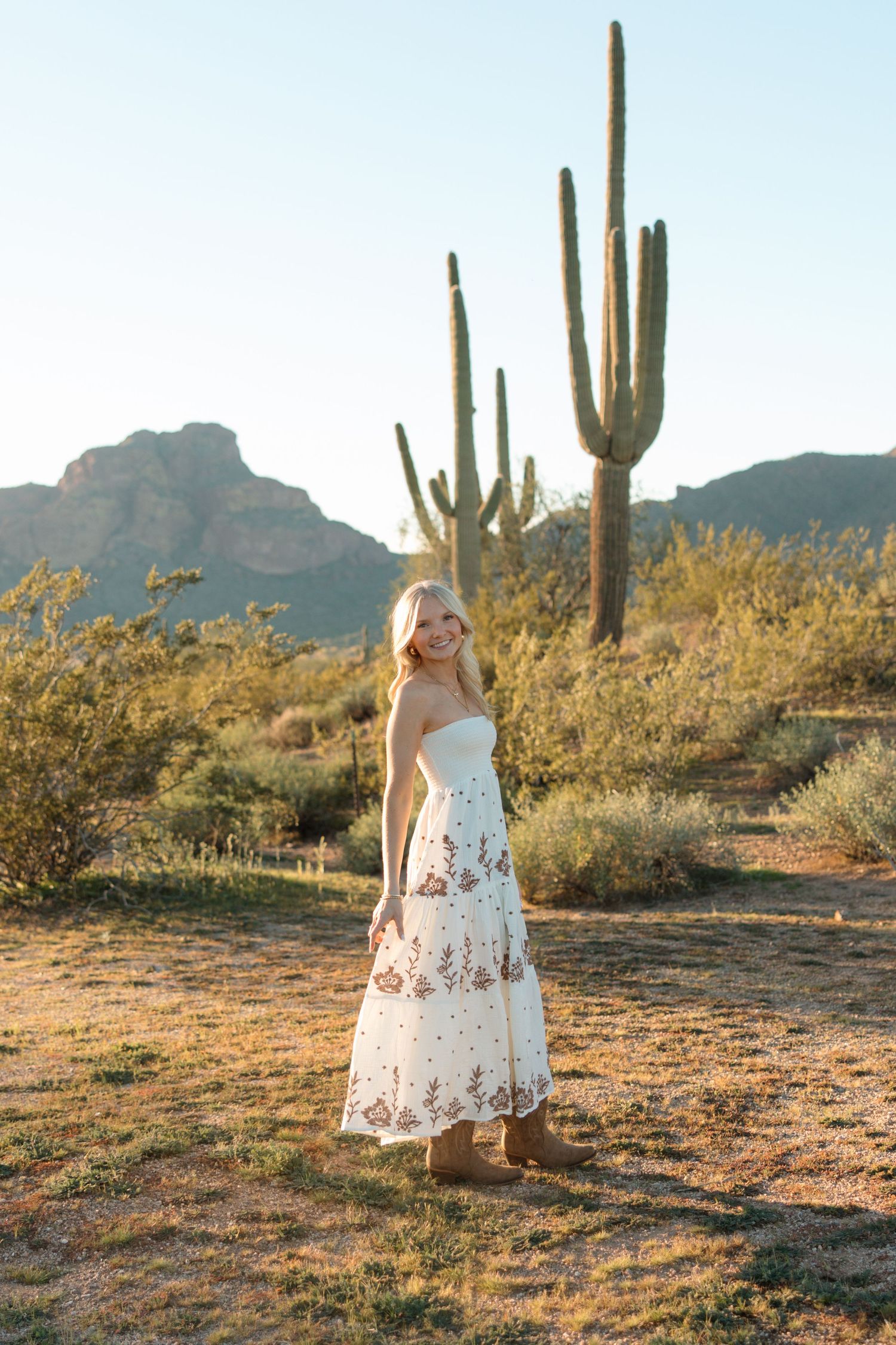 Blonde woman in white floral maxi dress and boots posing in Arizona desert with saguaro cacti at golden hour.