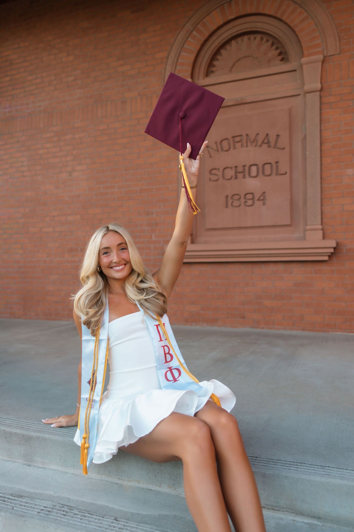 Smiling blonde graduate in white dress holds maroon cap at Normal School 1884 building wearing Pi Beta Phi sash.