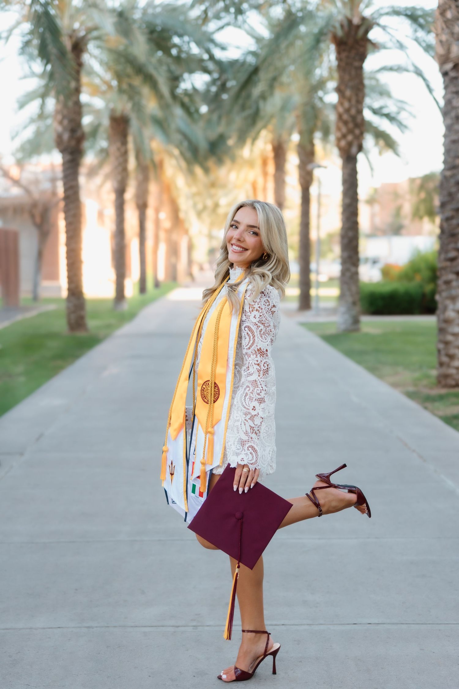 Smiling blonde graduate in white lace top holding maroon cap with gold stole on palm tree-lined campus path.