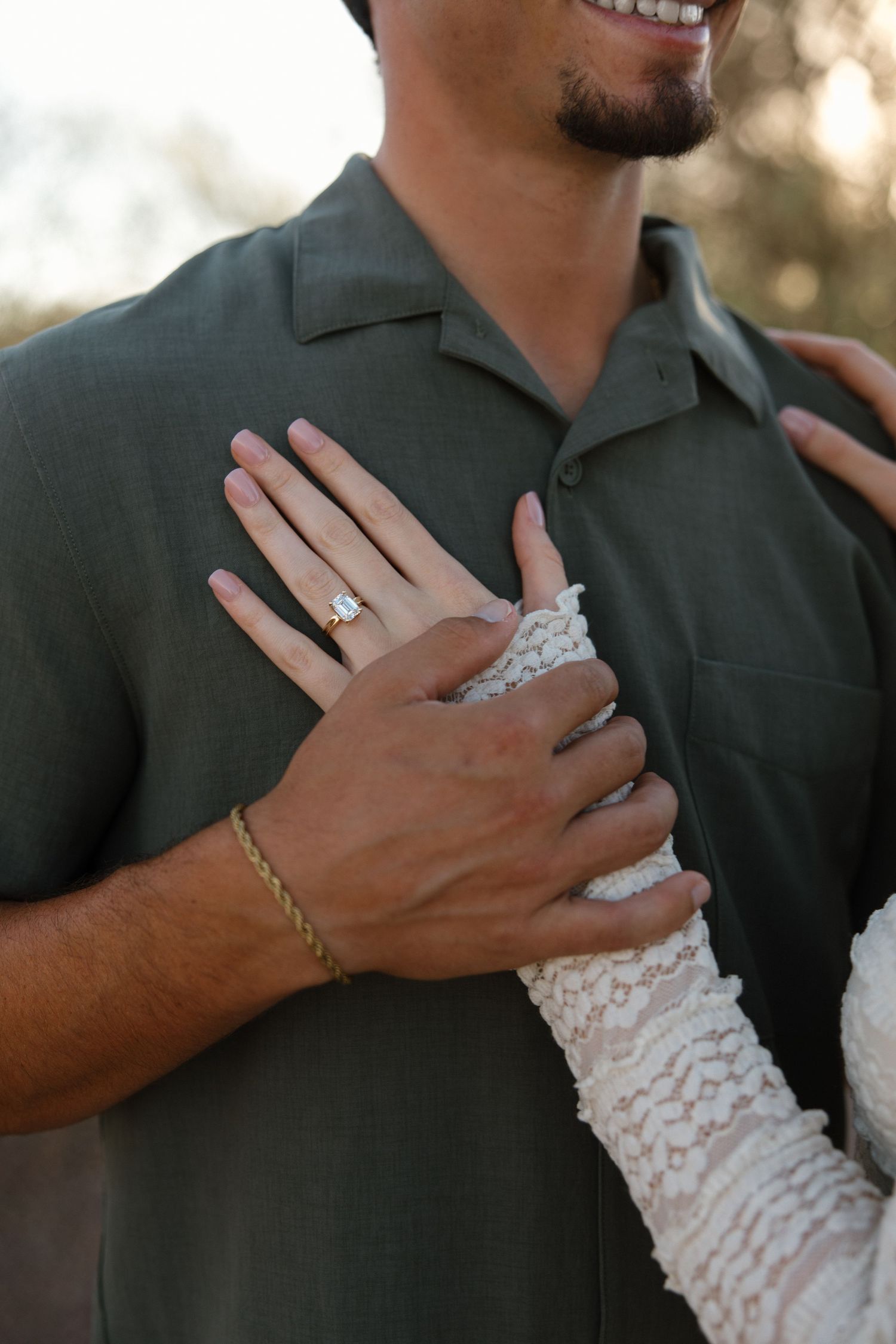 Close-up of a woman's hand with an engagement ring resting on a man's chest, both dressed elegantly outdoors.
