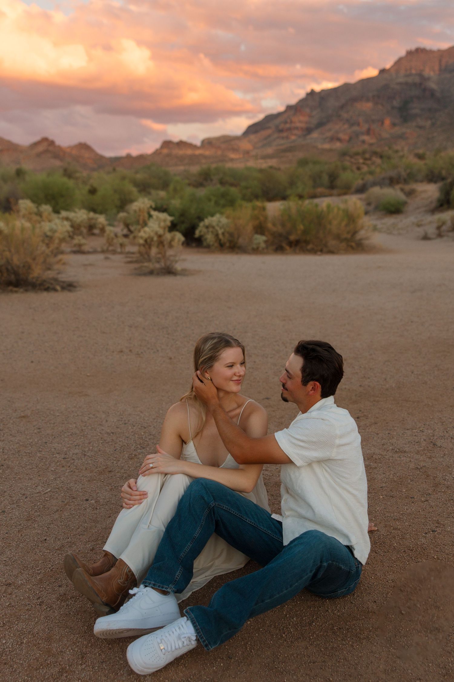Couple sitting on desert sand at sunset, man tenderly touching woman's face, mountains and pink sky behind them.