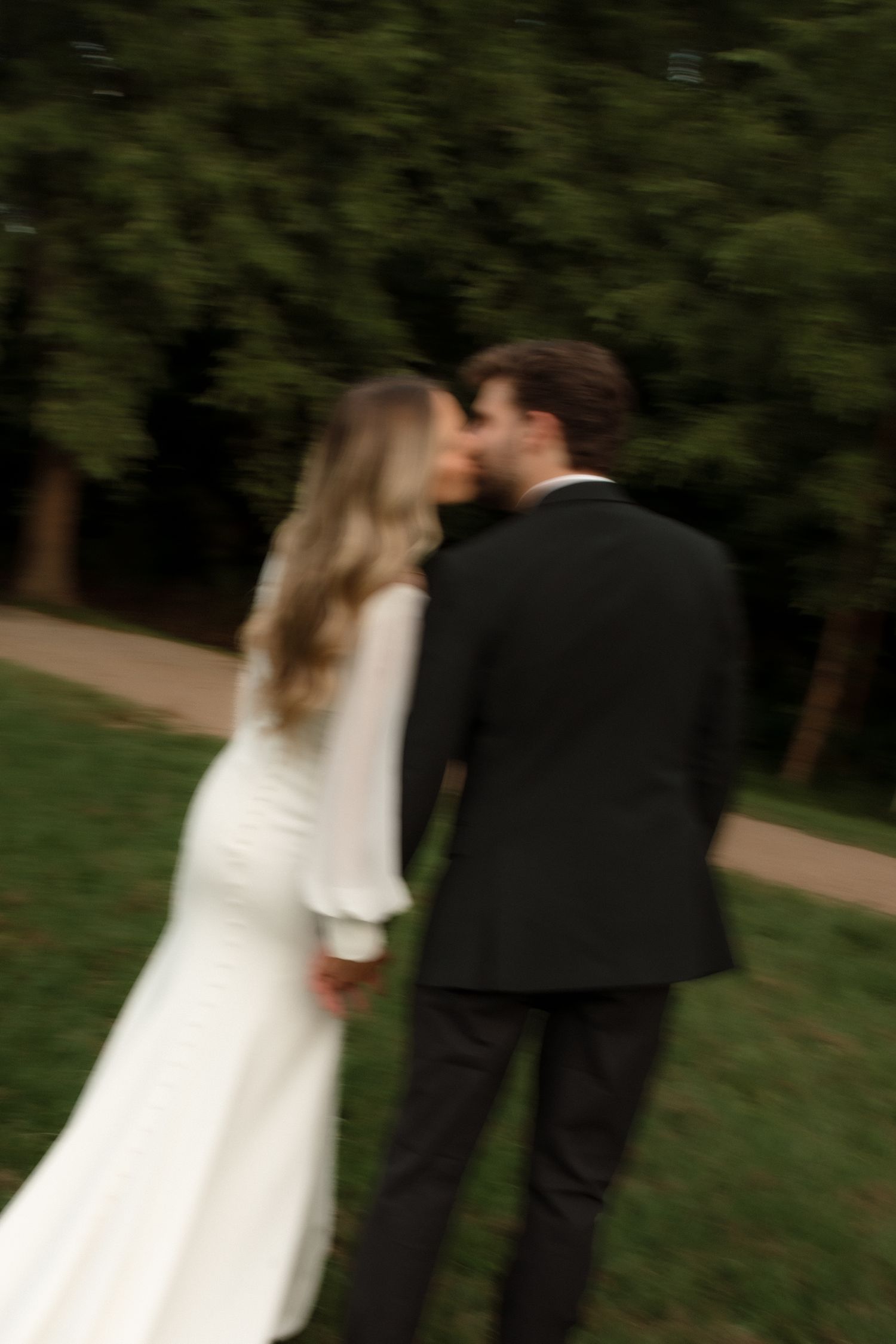 Bride and groom share a romantic kiss while holding hands in a lush green outdoor setting.