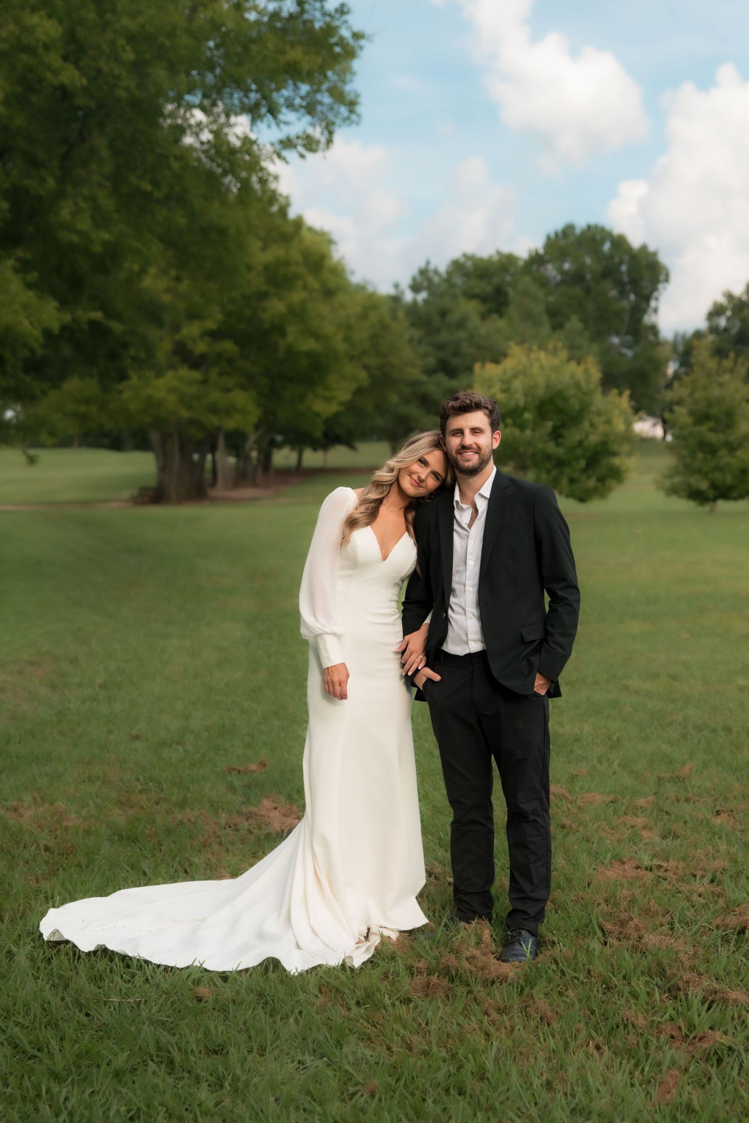 Bride in white gown with train and groom in black suit smiling together on green lawn with trees.