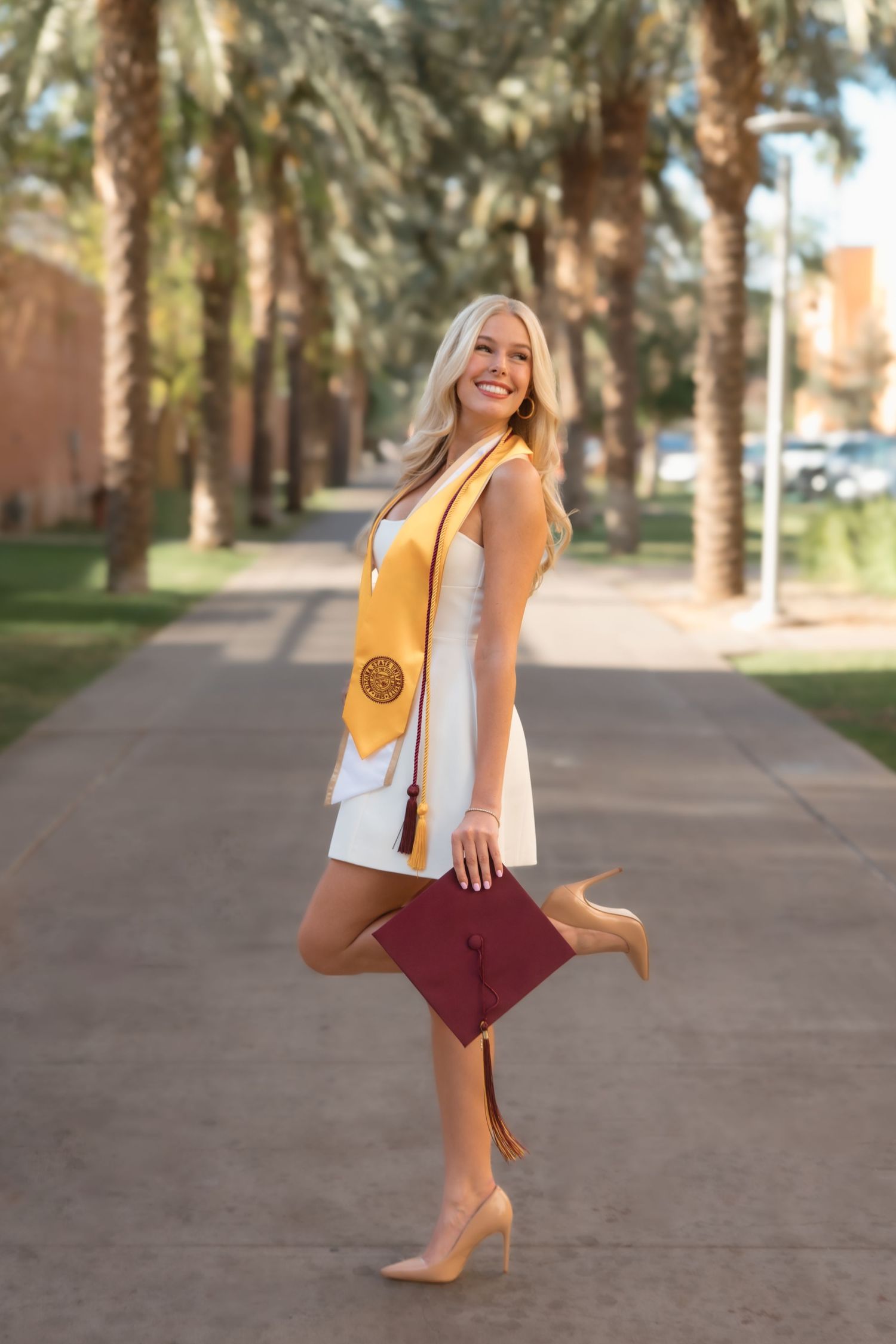 Smiling blonde graduate in white dress with gold ASU stole holds maroon cap among palm trees.