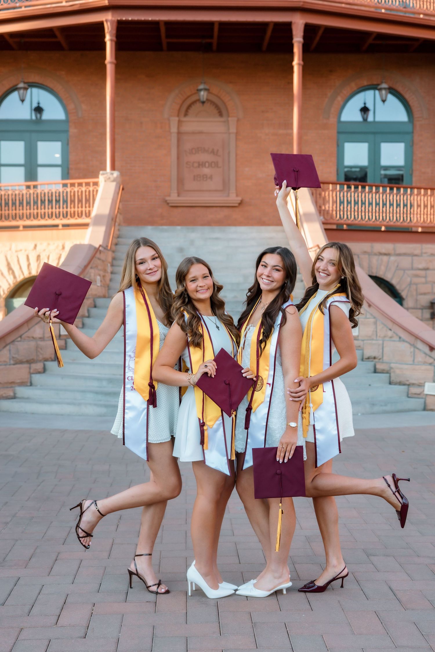 Four smiling female graduates in white dresses with gold sashes hold maroon caps at Arizona State University.