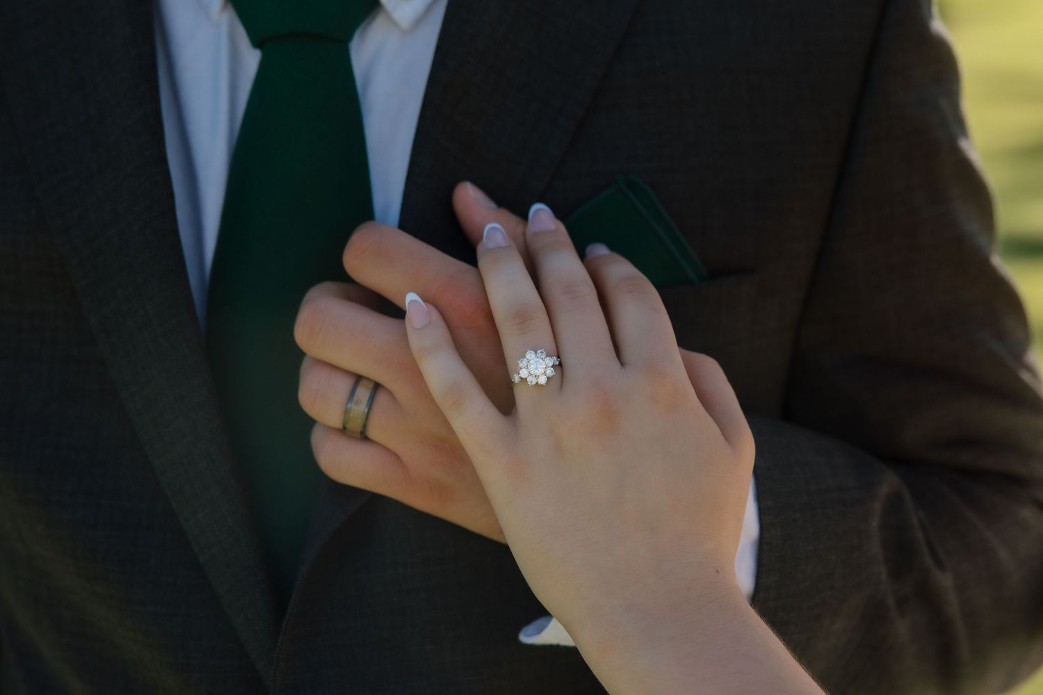 Close-up of bride and groom's hands showing diamond engagement ring and wedding bands, green tie visible.