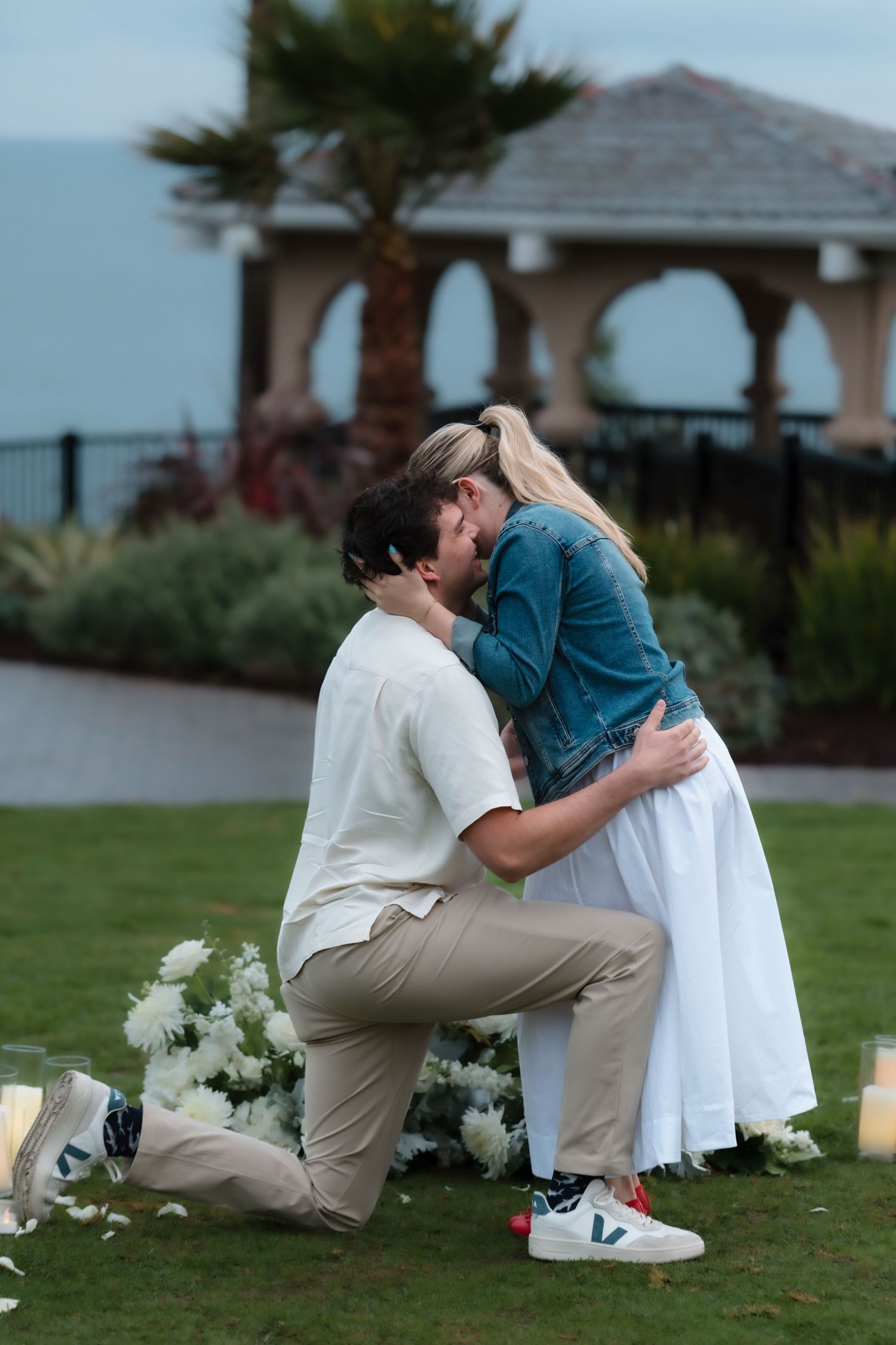 Couple sharing a kiss during outdoor marriage proposal surrounded by white floral arrangements and candles at dusk.