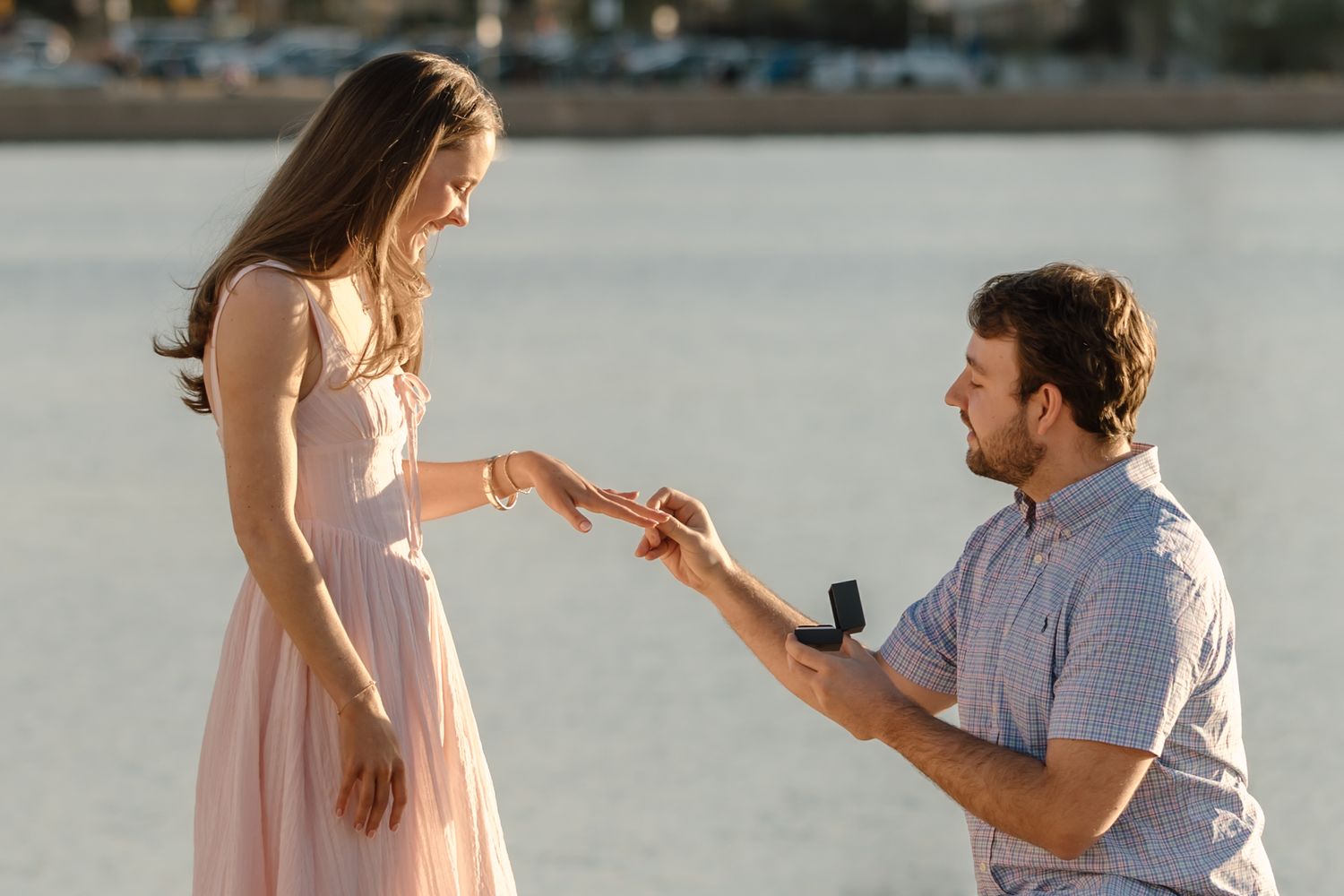 Man proposes to woman with ring box by waterfront, she reaches out her hand in a romantic outdoor setting.