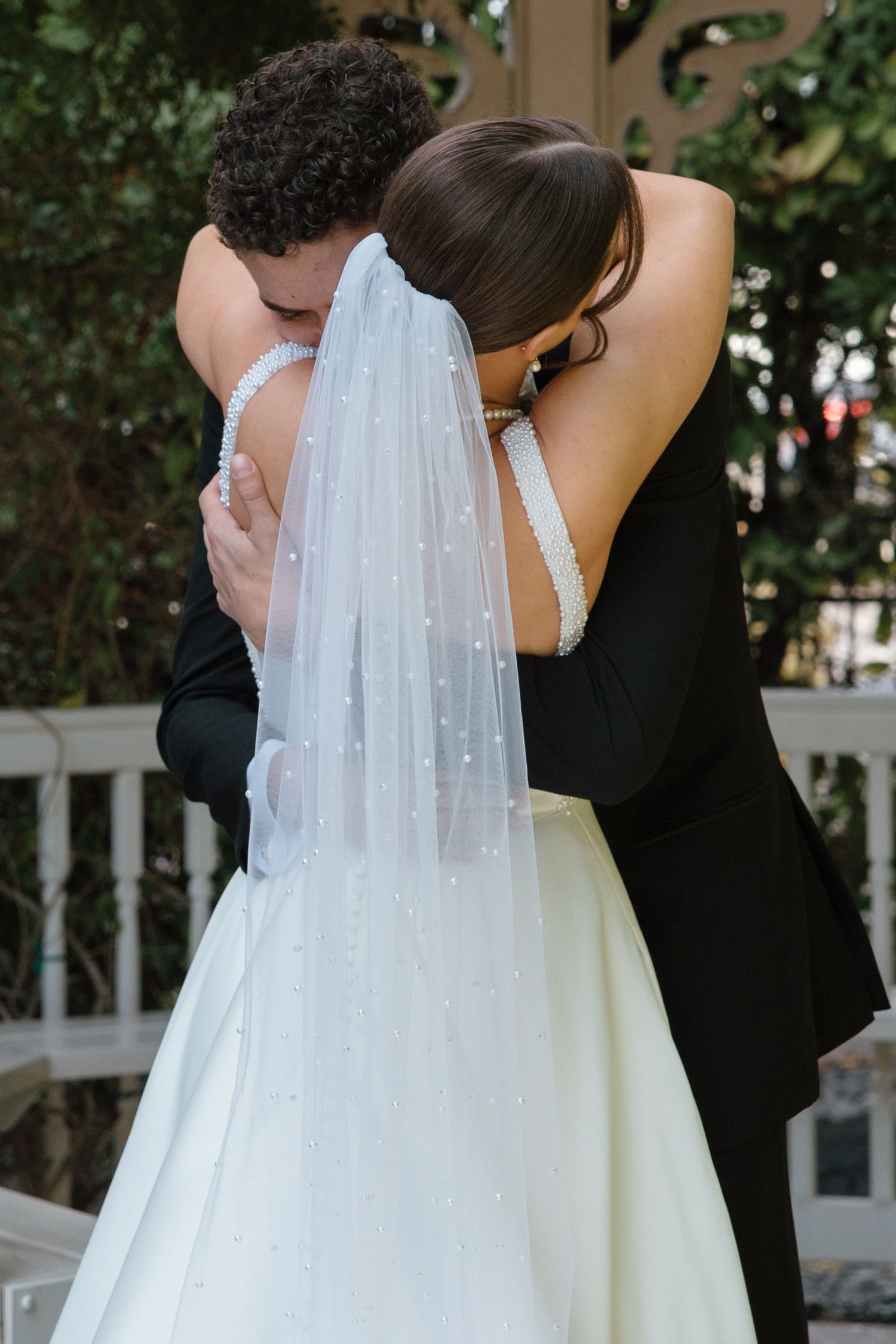 Bride and groom share emotional first look hug, bride wearing pearl-adorned veil at outdoor gazebo.