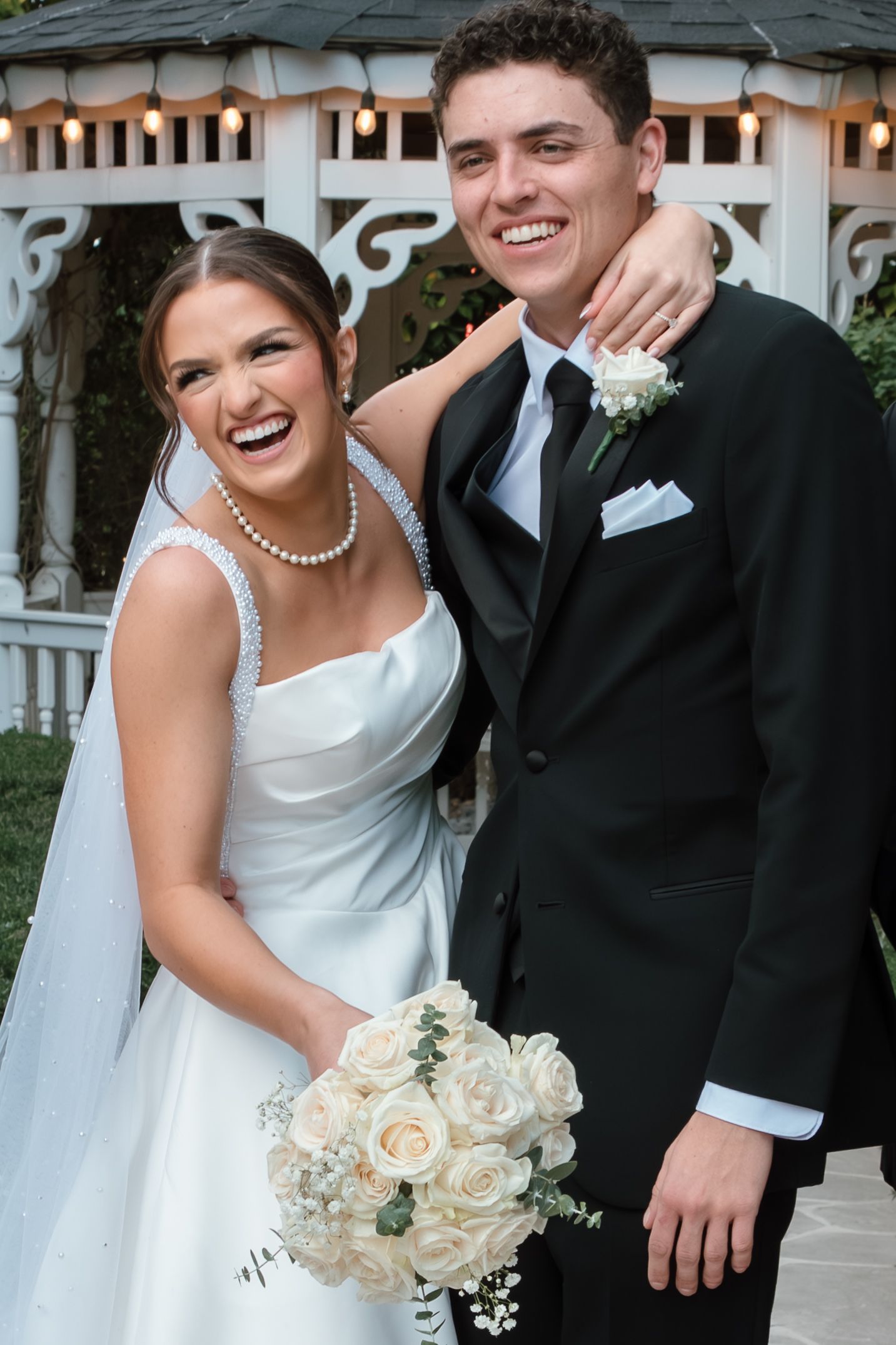 Joyful bride in white gown with pearl necklace and groom in black tuxedo posing together at outdoor wedding venue.