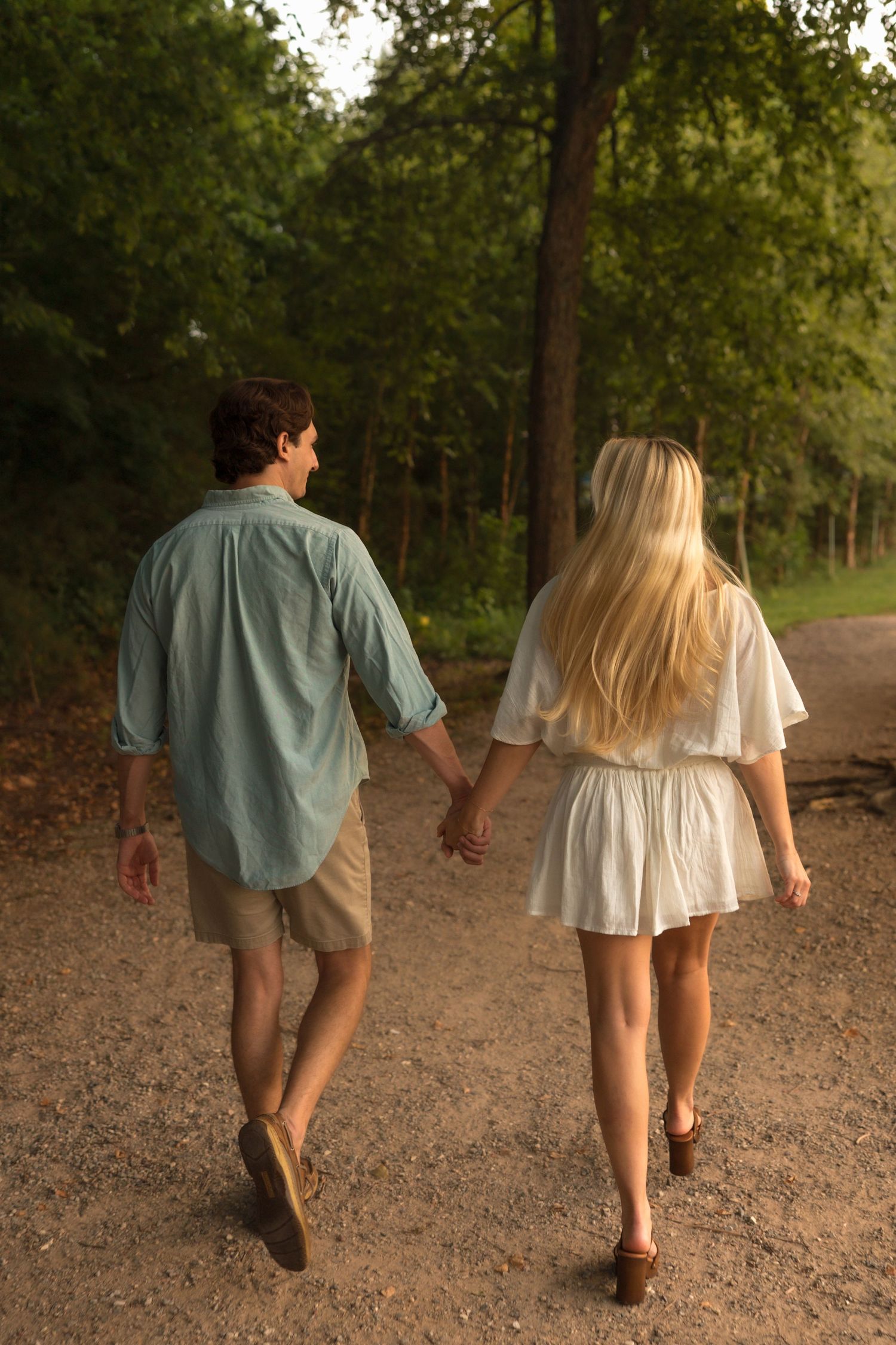 A couple walks hand in hand down a tree-lined gravel path during a warm sunset.