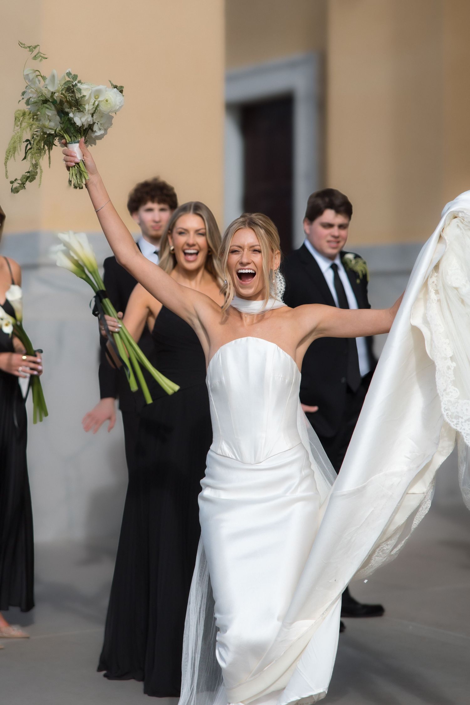 Joyful bride in strapless white gown celebrates outside church with wedding party in black attire.