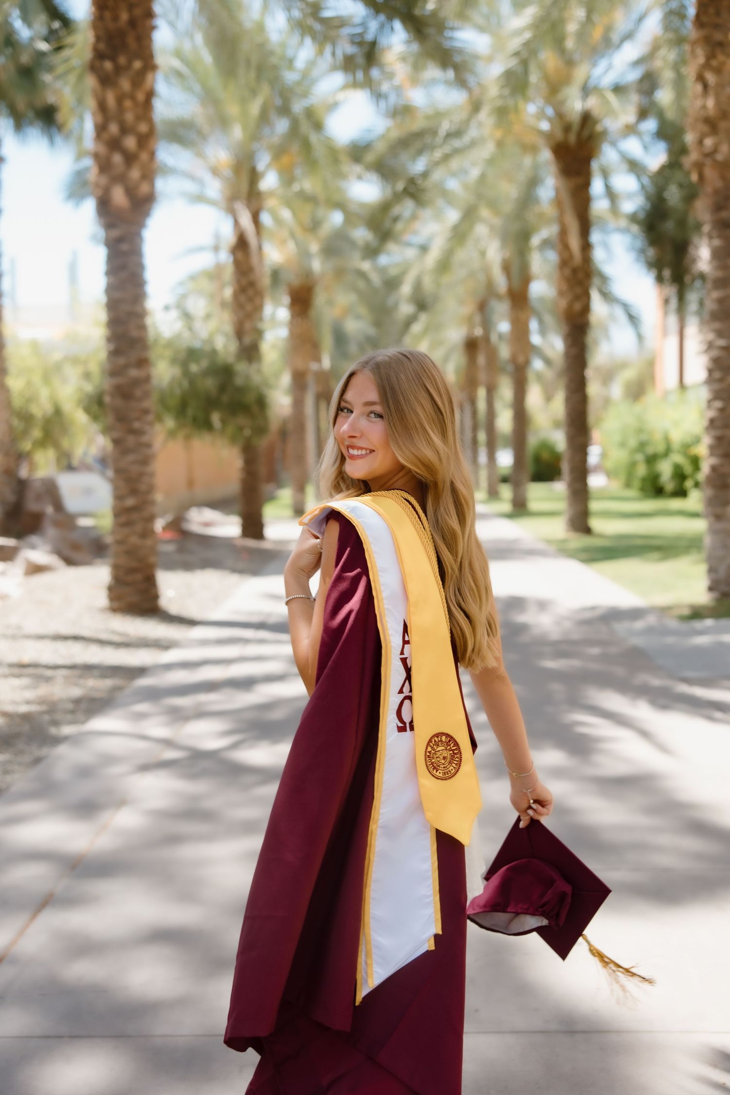 Smiling graduate in maroon gown and gold sash holds cap among palm trees on sunny campus walkway.