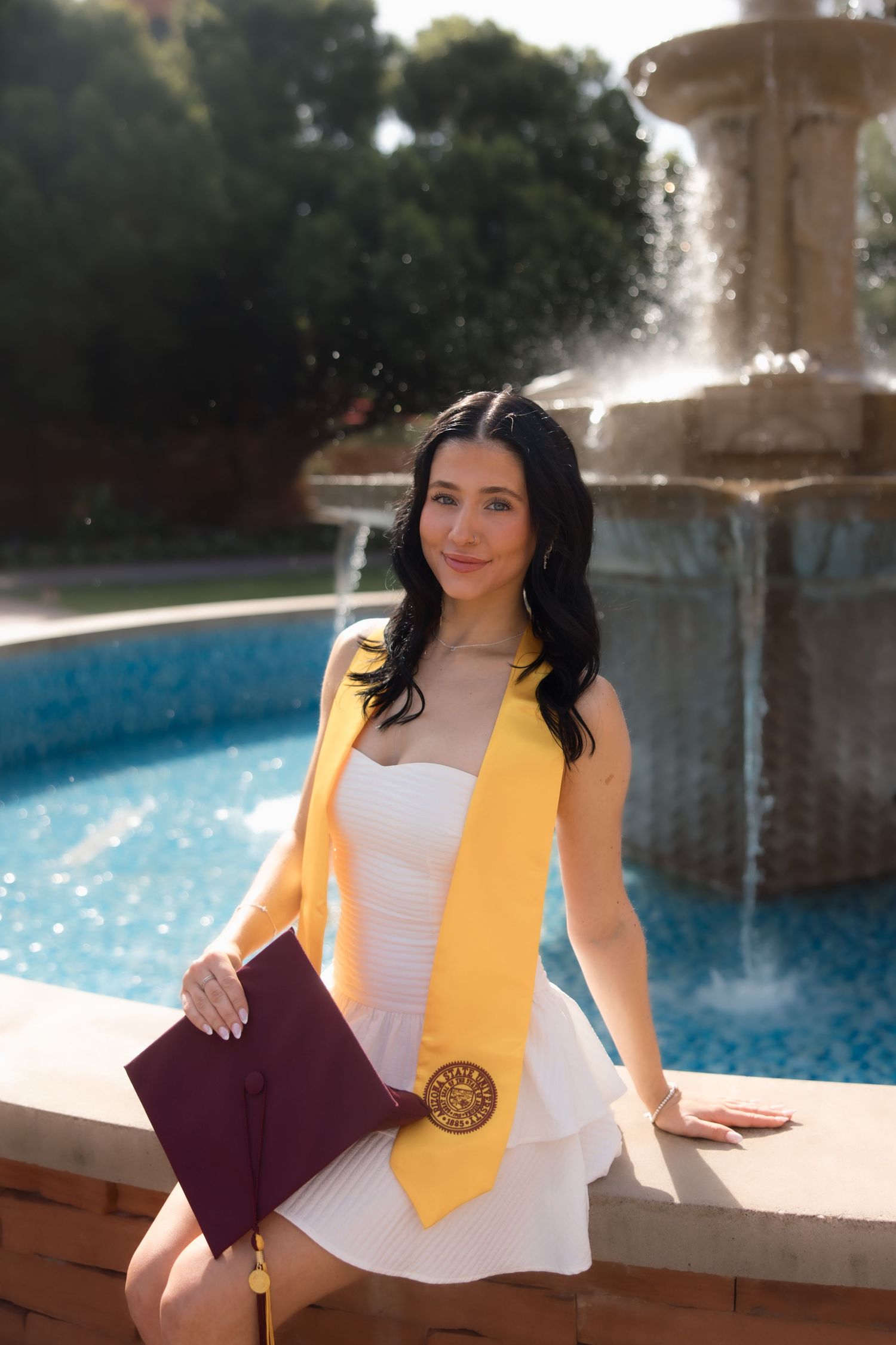 Young woman in white dress with gold graduation stole holds maroon cap by a fountain outdoors.