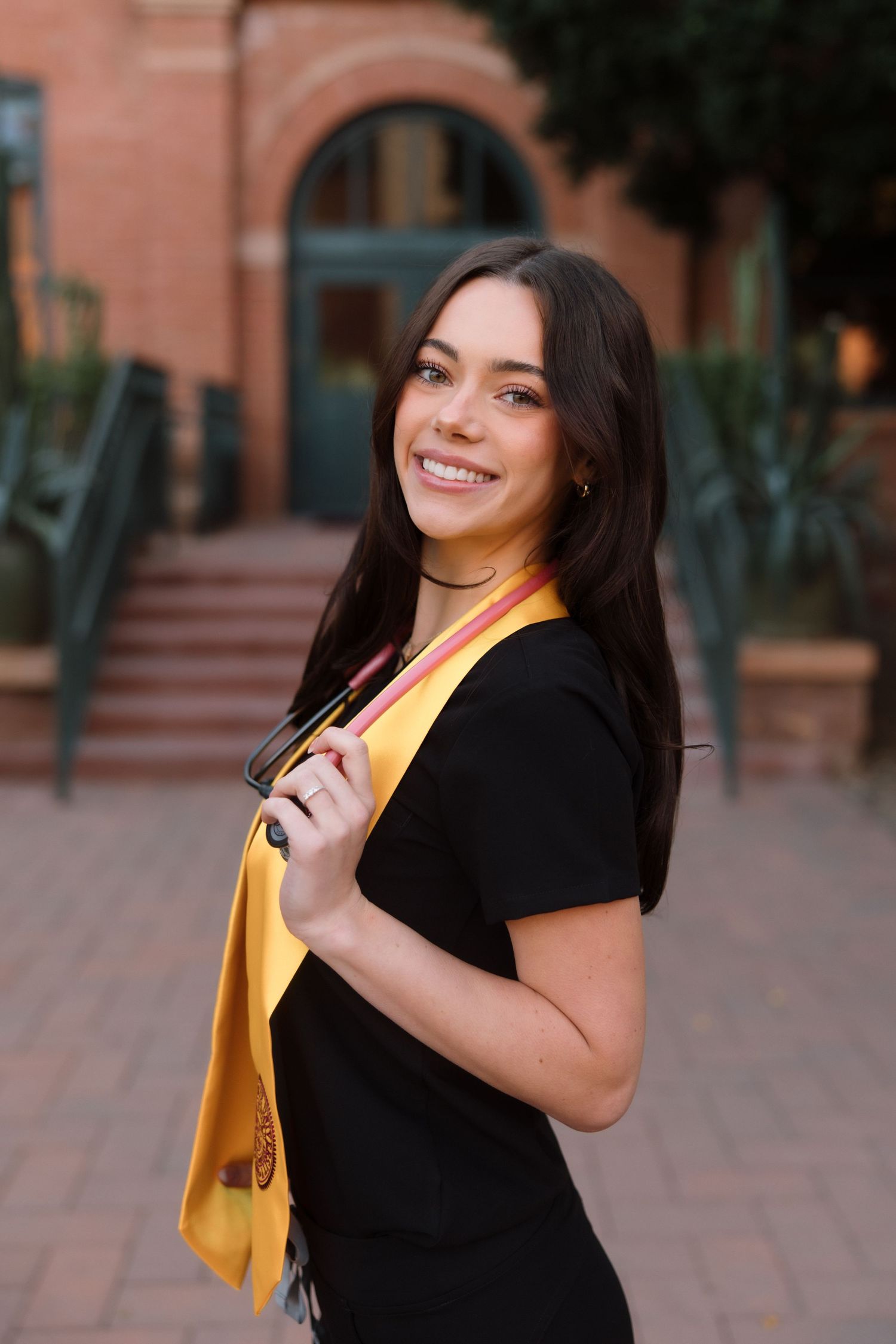 Brunette female graduate smiling in black dress with gold graduation stole on university campus.