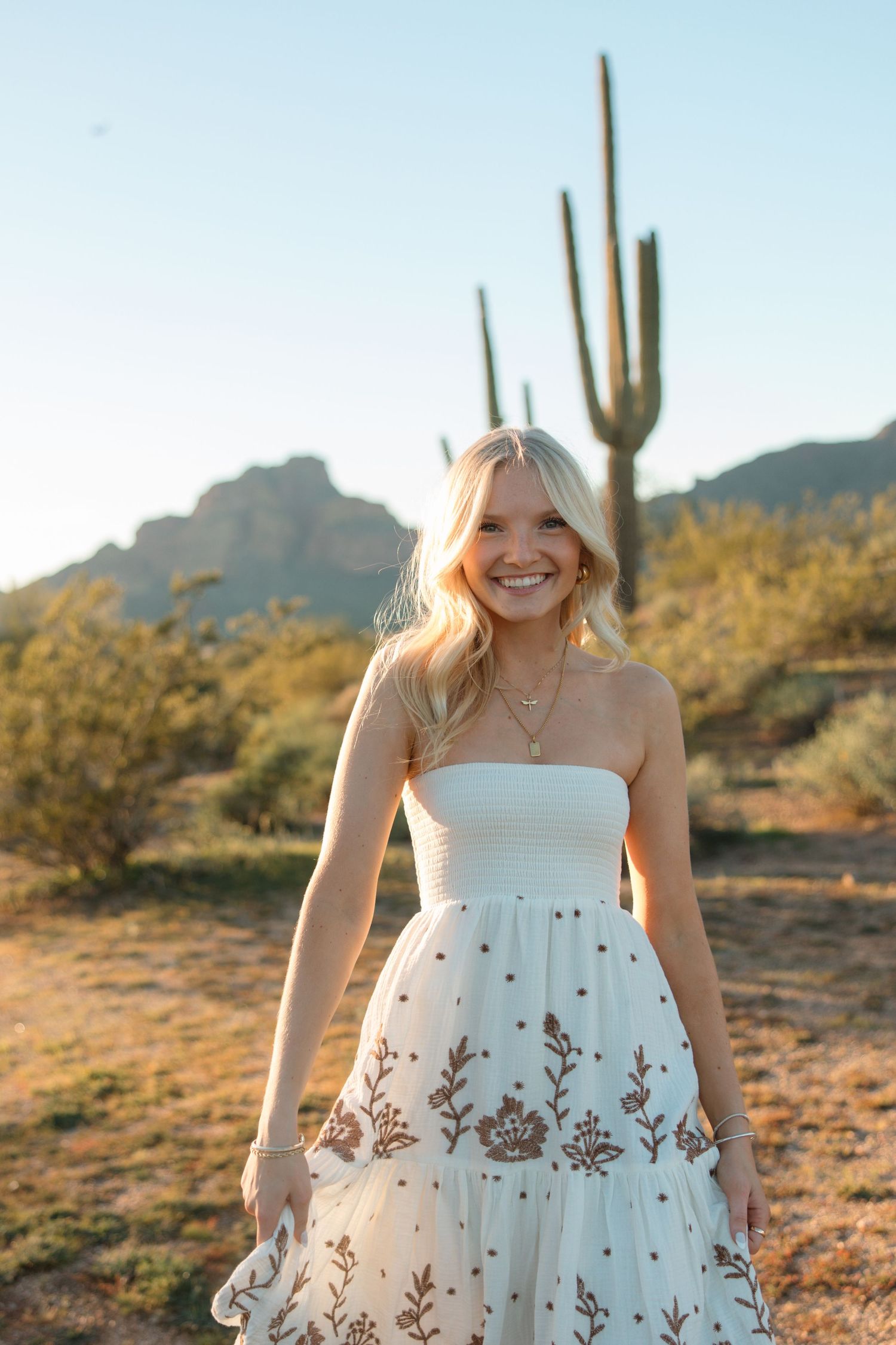 Happy blonde woman in white embroidered strapless dress standing in sunny desert landscape.