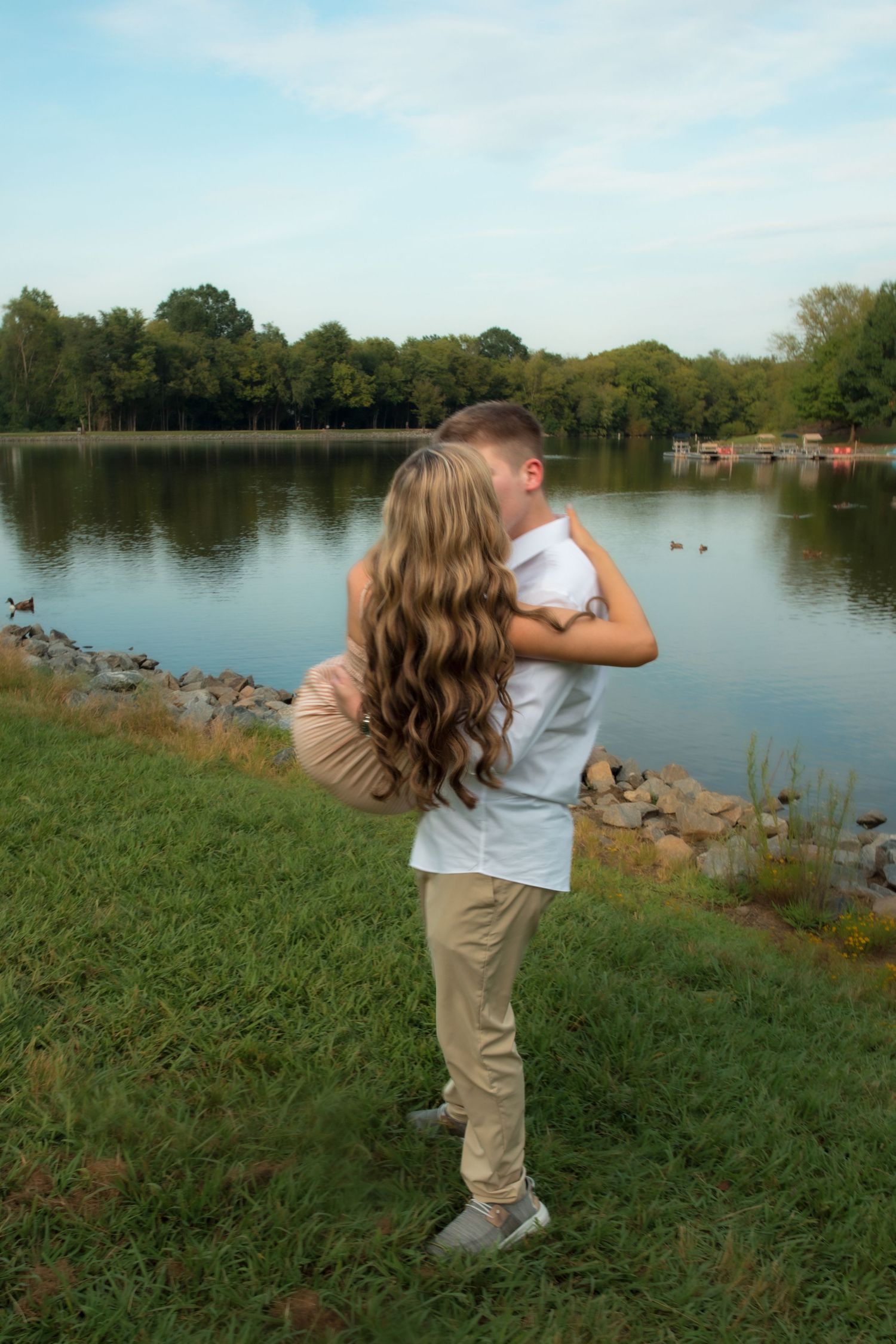 Couple embracing by a serene lake surrounded by lush green trees and rocky shoreline on a sunny day.