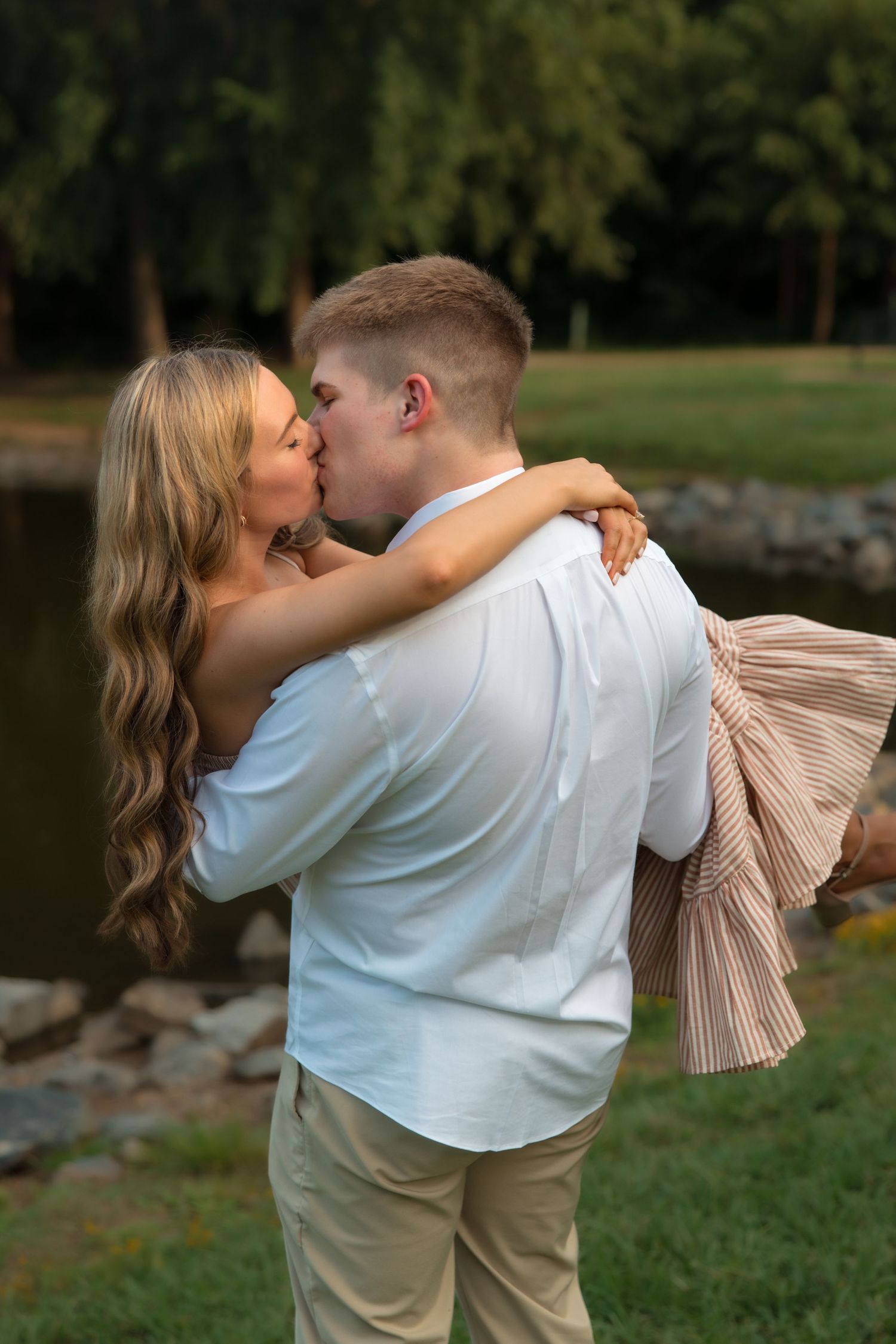 A man lifts and kisses a woman with long blonde hair outdoors near a pond, both dressed in casual elegant attire.