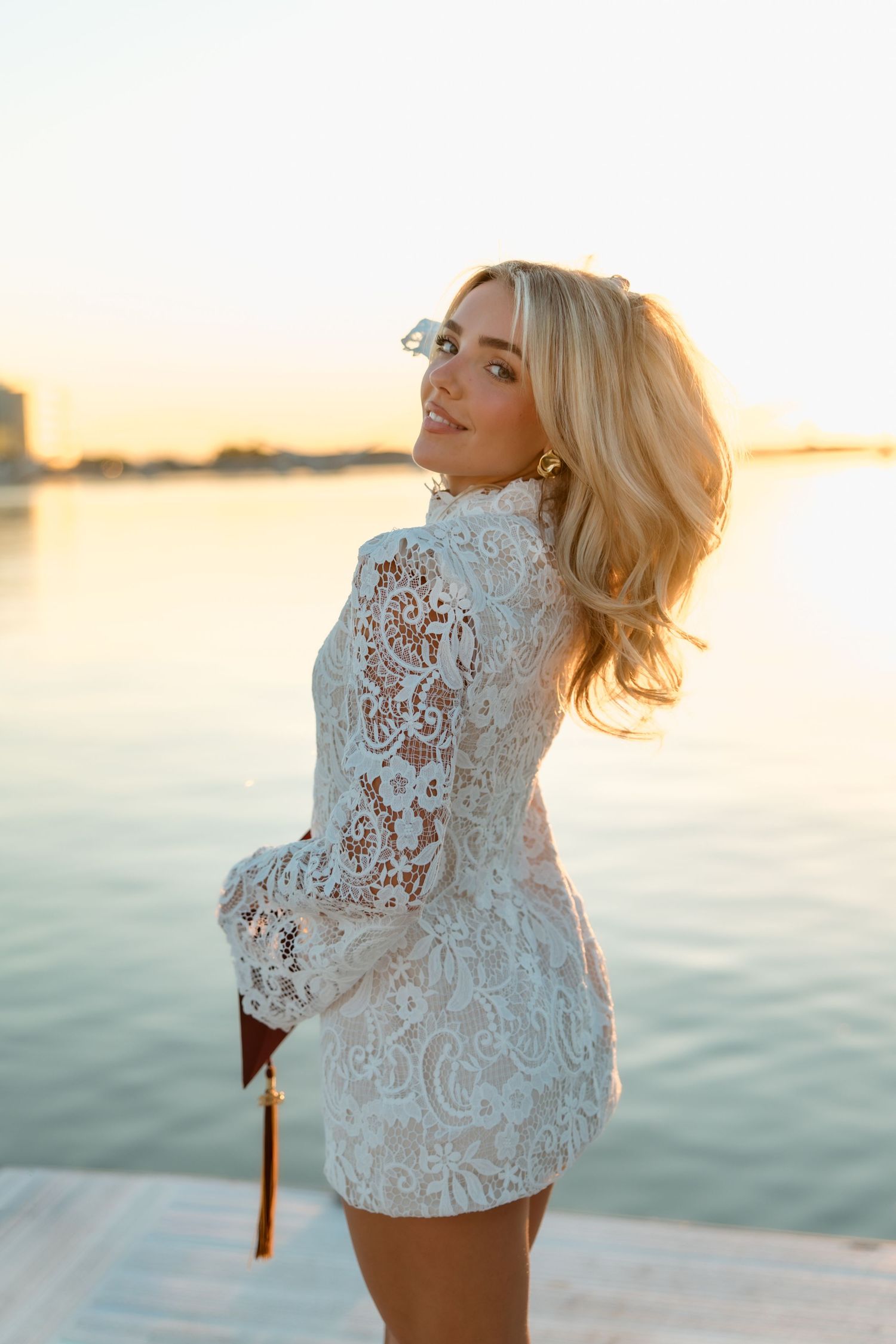 Blonde woman in white lace mini dress smiling over shoulder by waterfront at golden hour sunset.