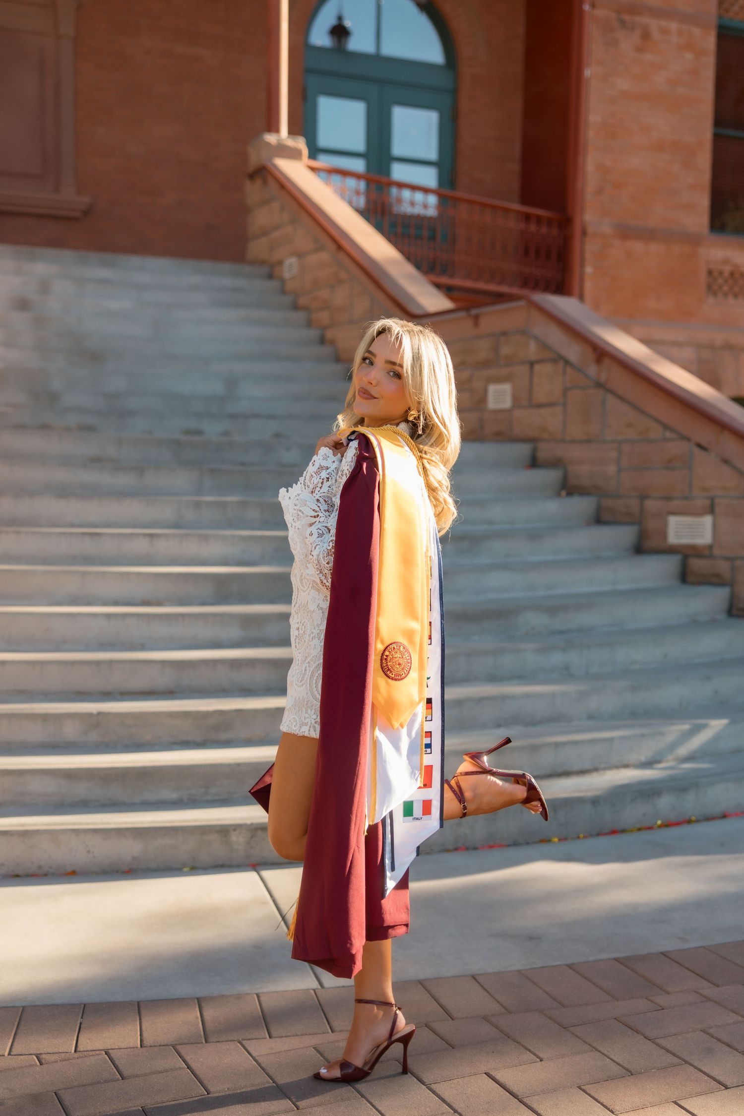 Blonde woman in white lace dress with maroon graduation gown and gold stole poses on brick steps.