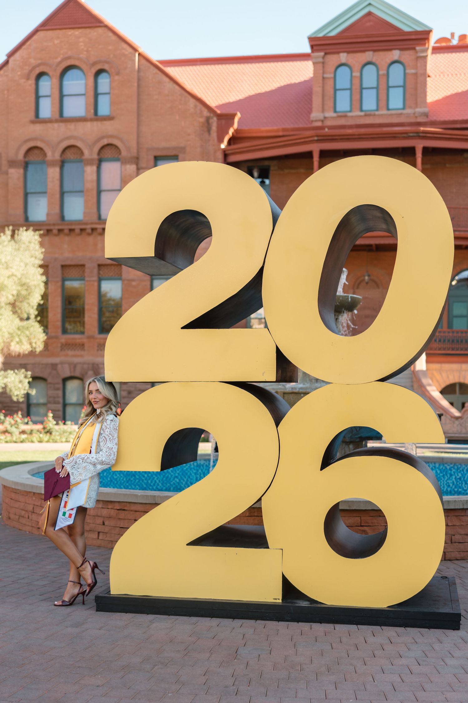 Woman posing beside large gold 2026 numbers sculpture on a university campus with a red brick building backdrop.