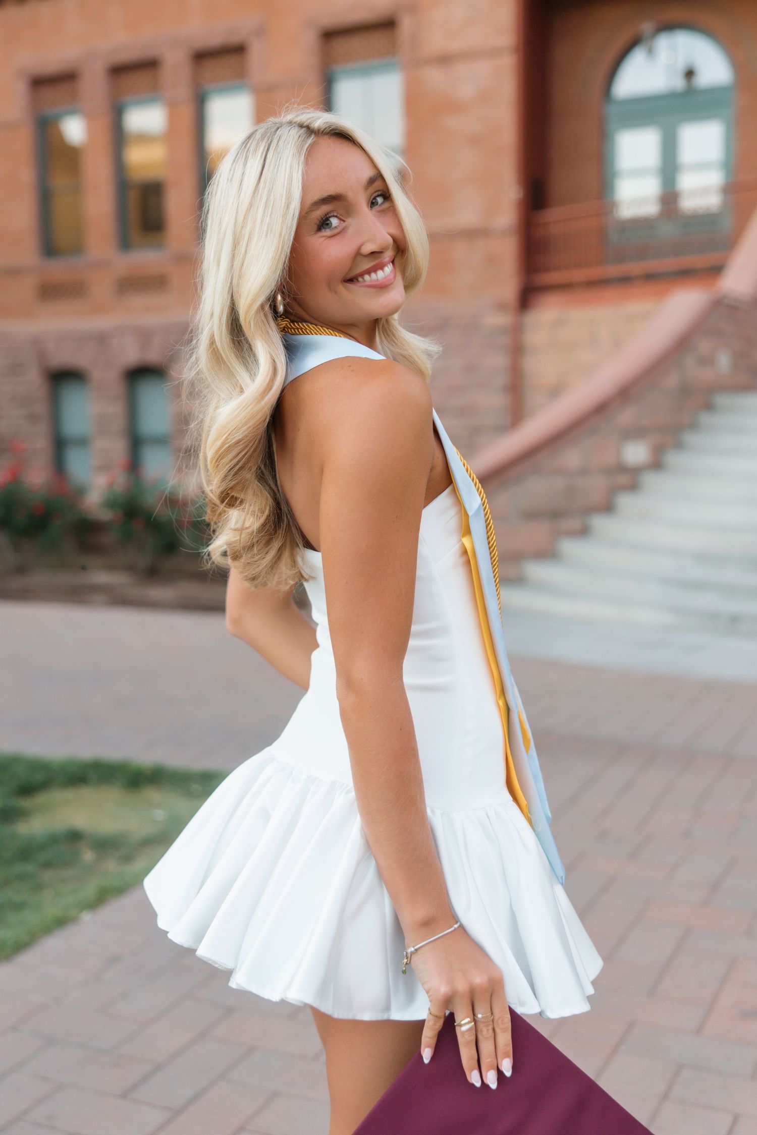 Smiling blonde woman in white dress holding maroon folder outdoors near brick building steps.