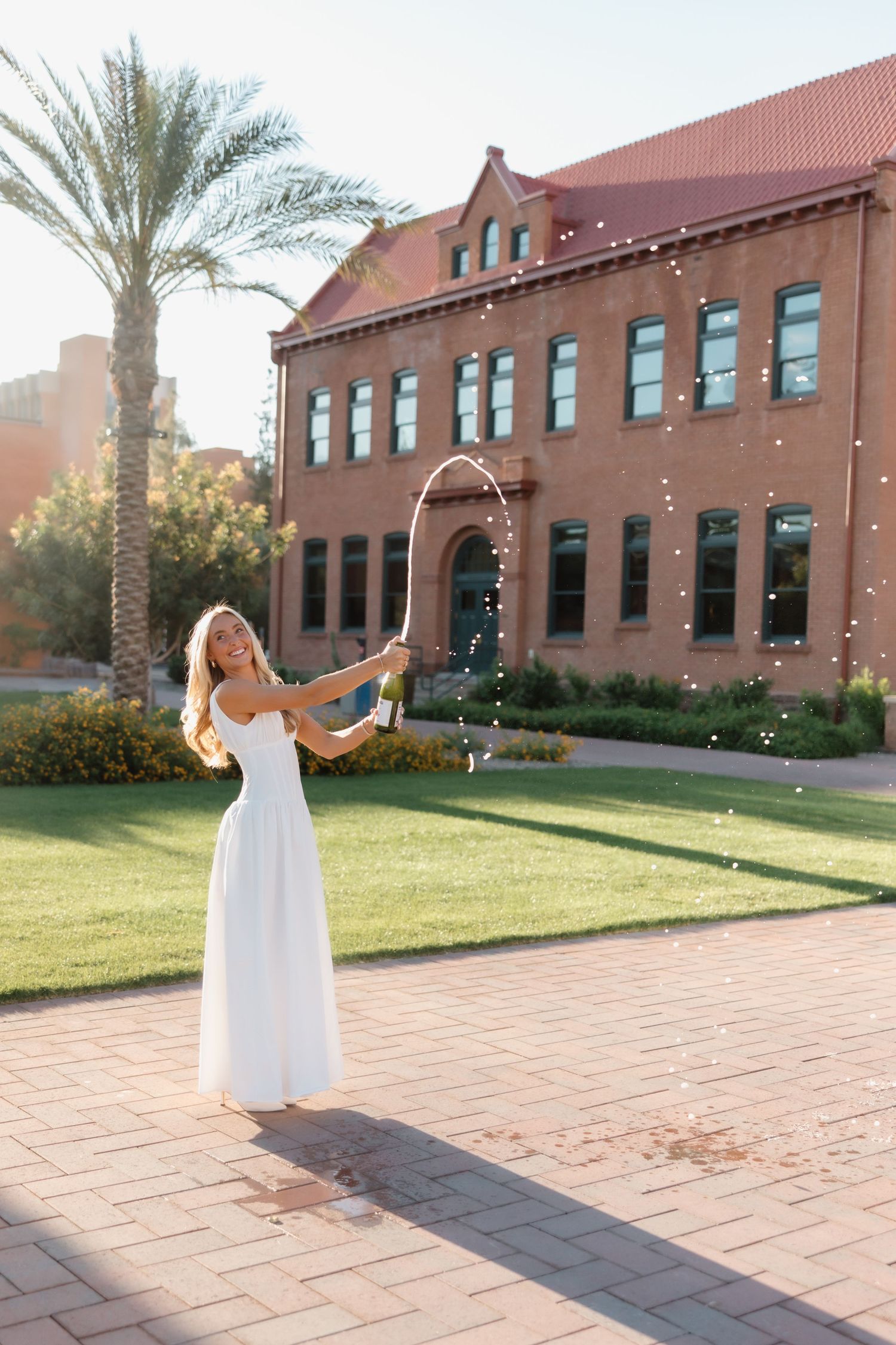 Joyful woman in elegant white dress spraying champagne outside historic brick building with palm tree.