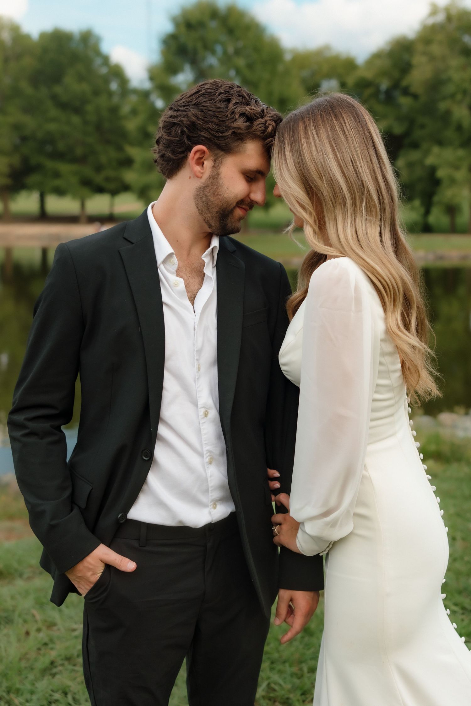 Bride and groom in black suit share an intimate moment near a serene pond surrounded by trees.