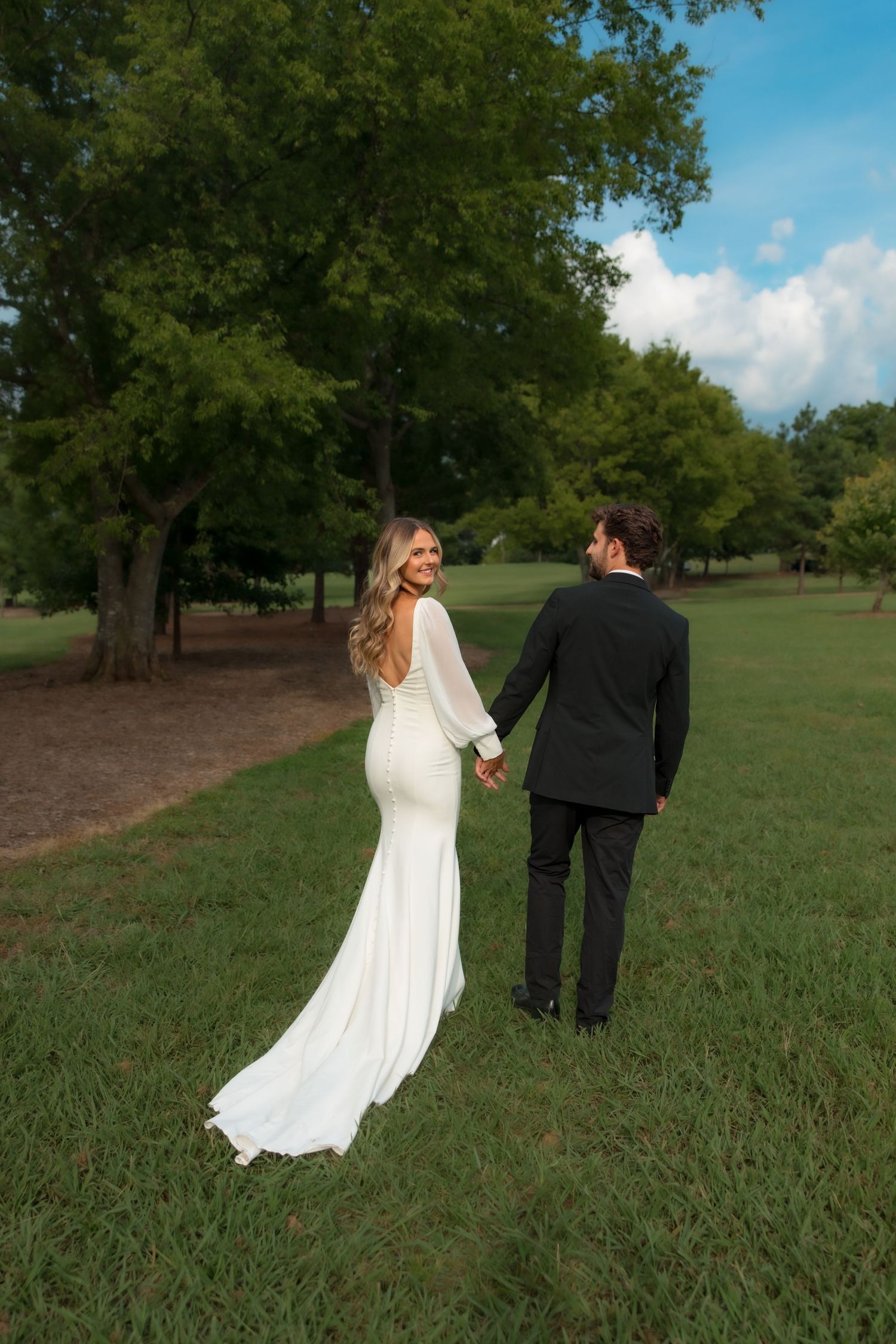 Bride and groom holding hands walking through lush green park in wedding attire.