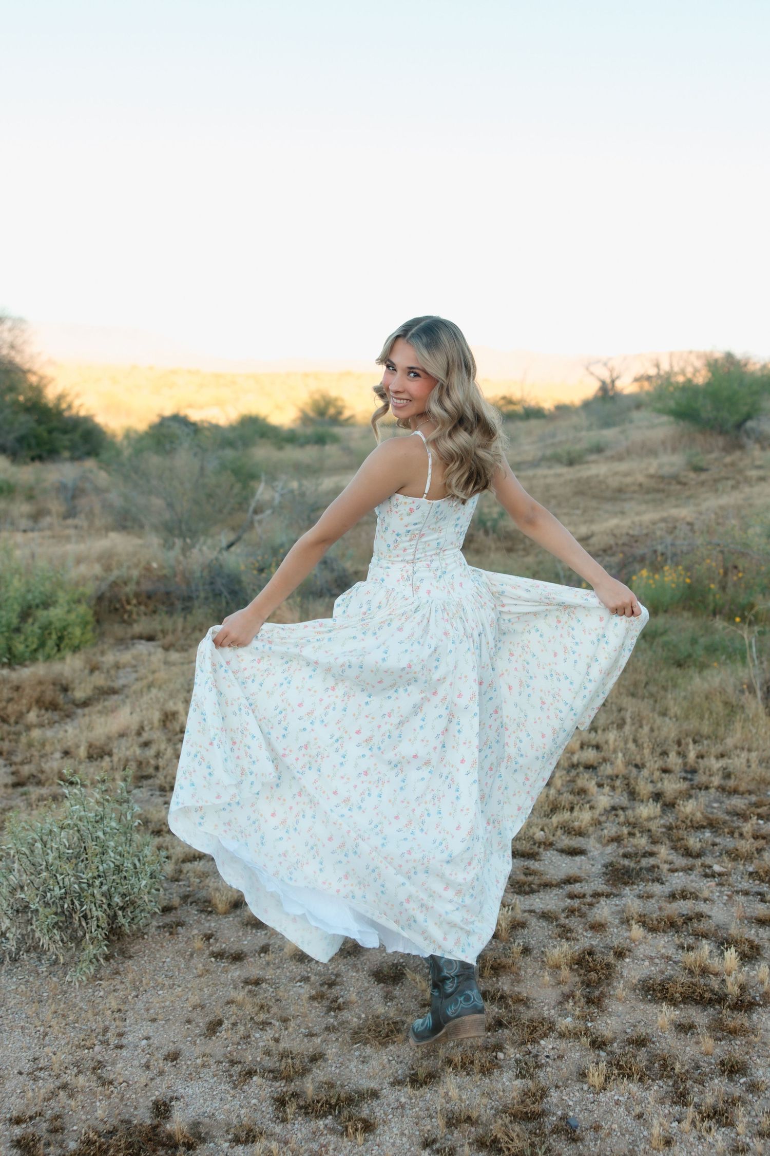 Young woman in white floral maxi dress twirling outdoors in desert landscape at golden hour.