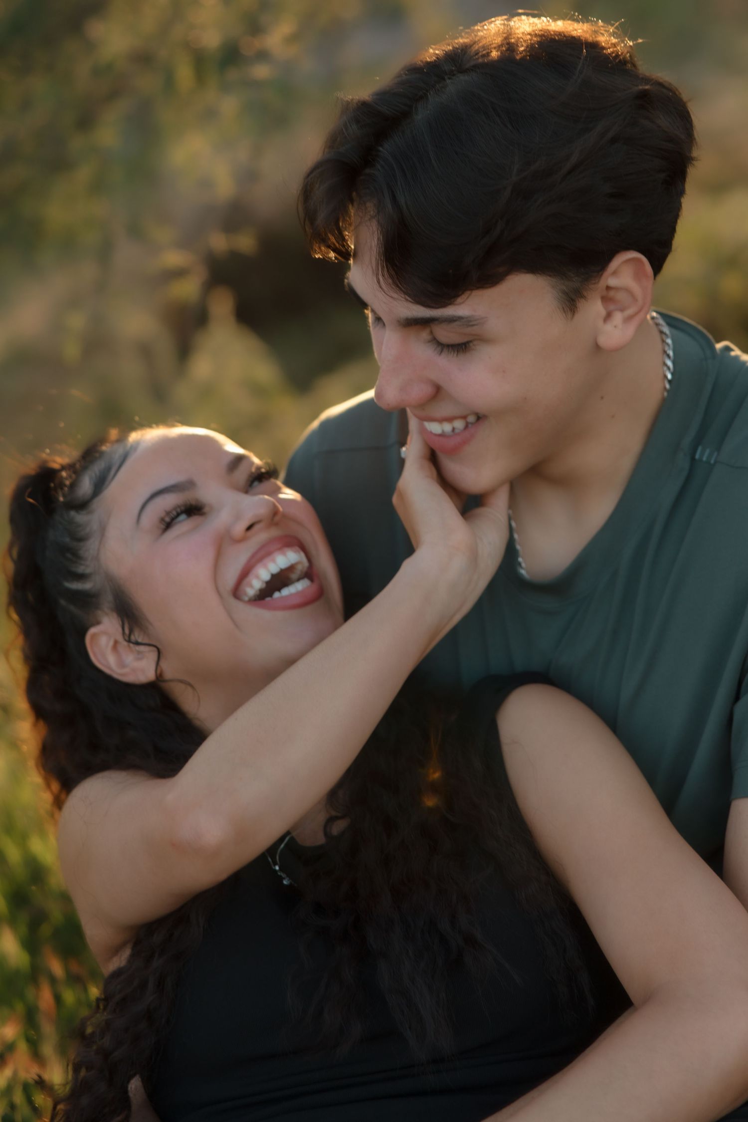 A laughing couple shares a joyful moment outdoors, the woman gently touching the man's smiling face.