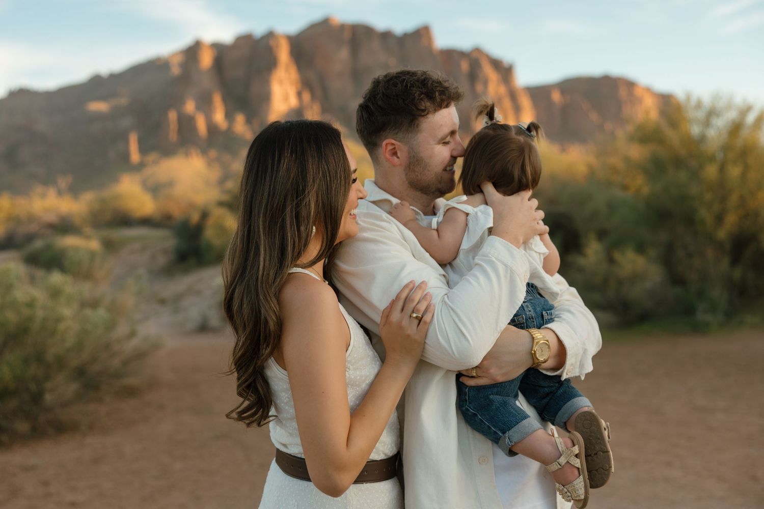 Parents sharing a tender moment kissing their young daughter in a warm desert landscape at sunset.