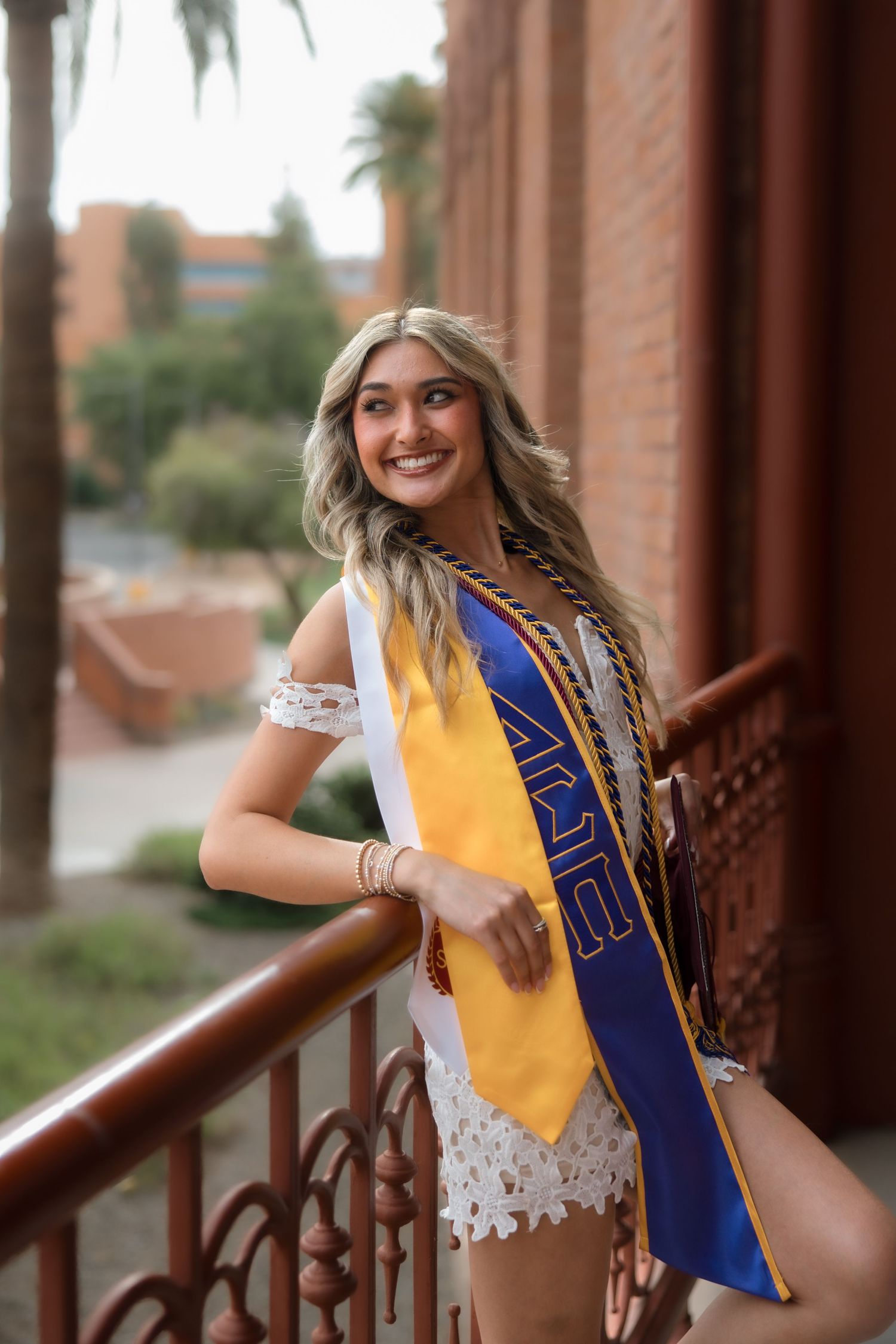 Smiling female graduate leans on railing wearing white lace outfit with Alpha Phi and gold ASU graduation stoles.