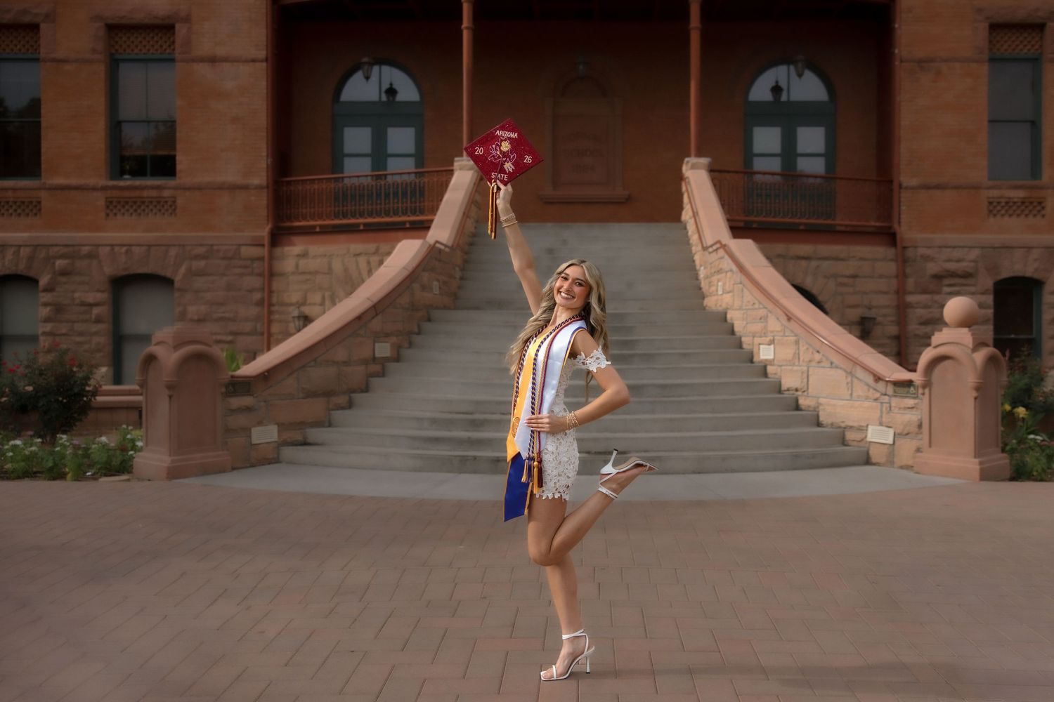 Smiling ASU graduate in white dress tossing maroon graduation cap in front of historic Old Main building staircase.