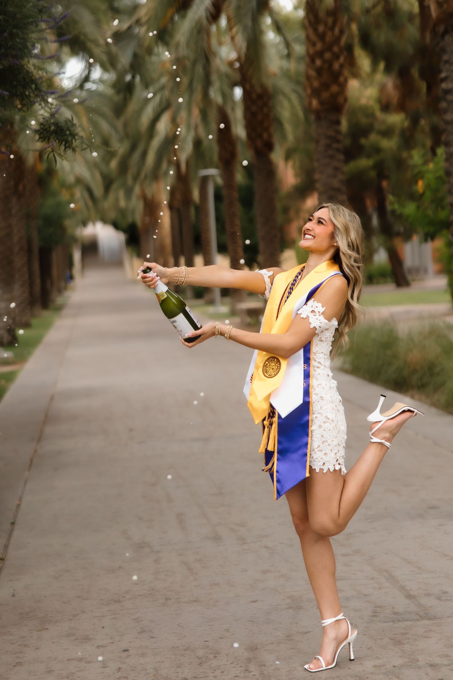 Joyful graduate pops champagne bottle wearing white lace dress with gold and blue graduation stoles among palm trees.