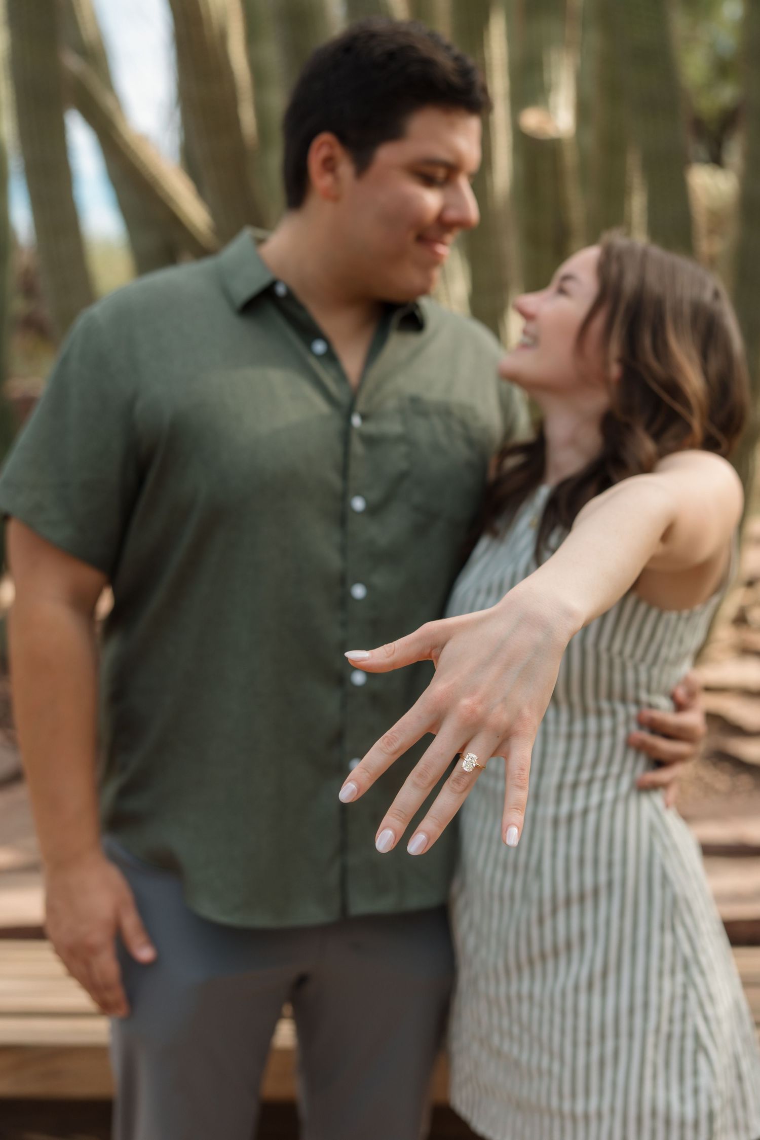 Newly engaged woman showing off diamond ring while embracing partner near desert cacti.