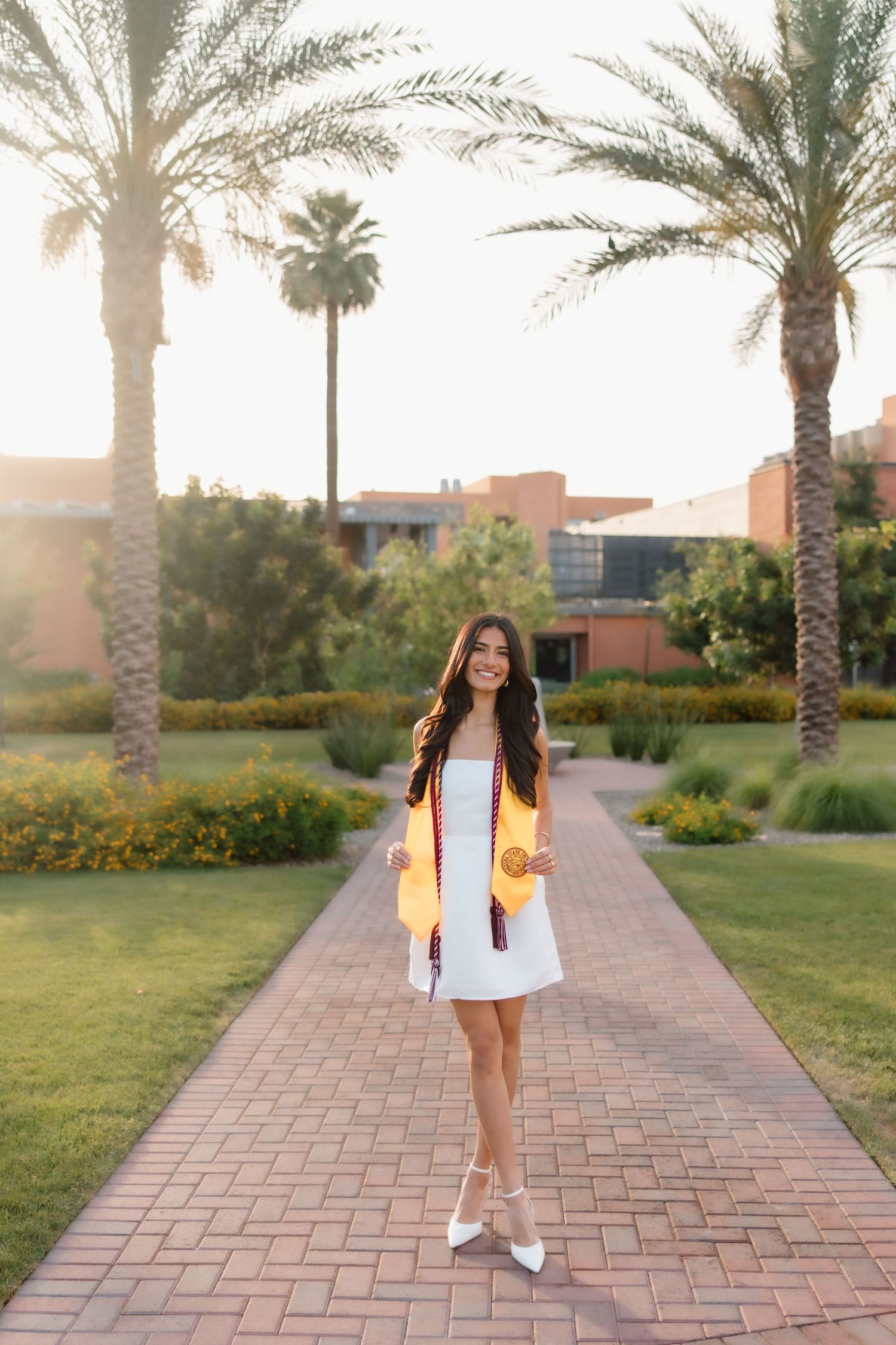 Smiling graduate in white dress and gold sash poses on brick path surrounded by palm trees at sunset.