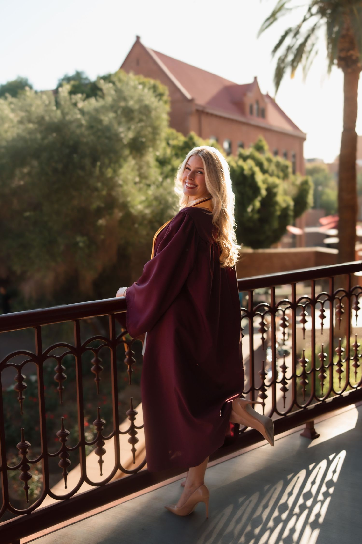 Blonde woman in maroon graduation gown smiles on ornate balcony with palm trees and brick building.