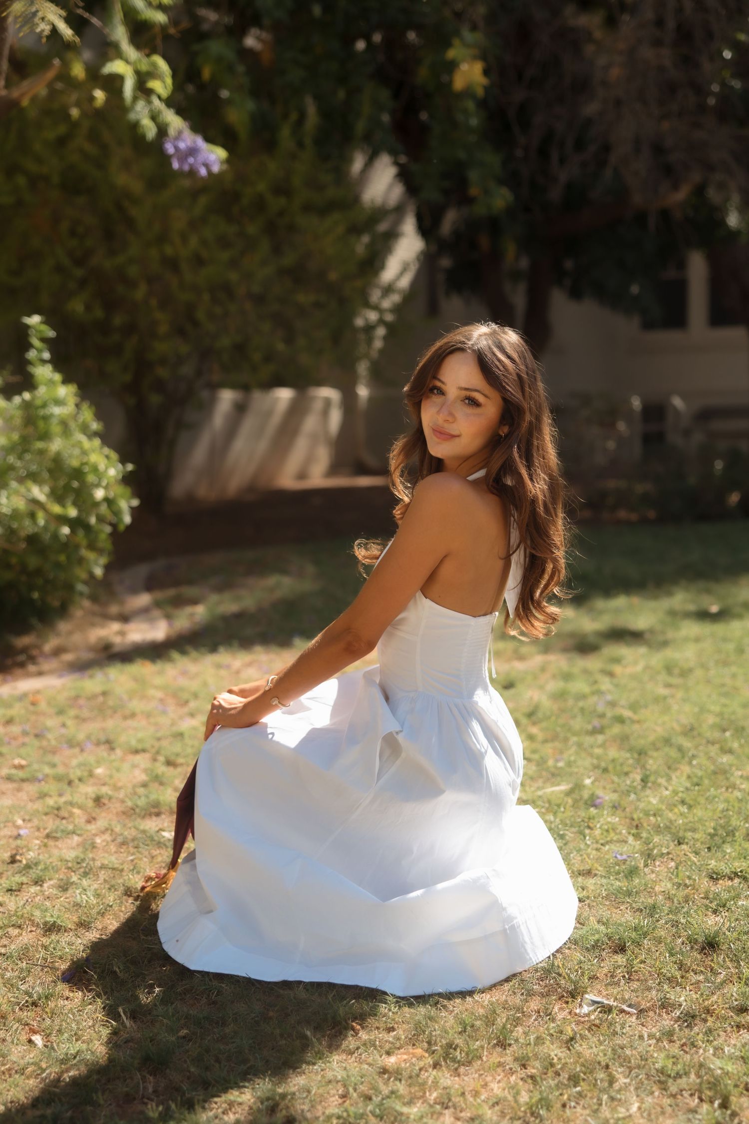 Young woman in white strapless dress sitting gracefully on green grass in a sunlit garden.