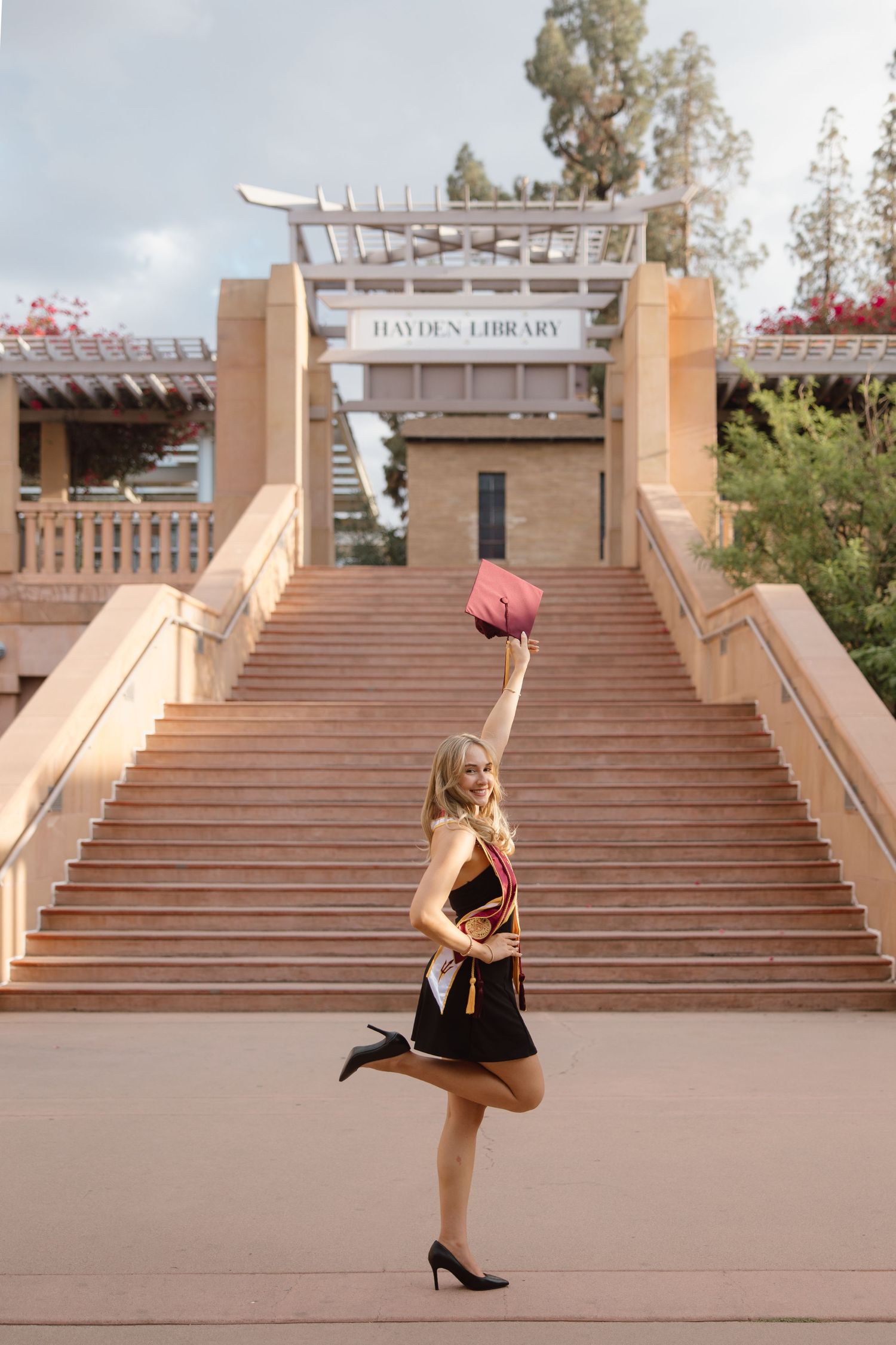 Smiling blonde graduate celebrates in front of Hayden Library steps, raising her cap while posing in black dress and heels.