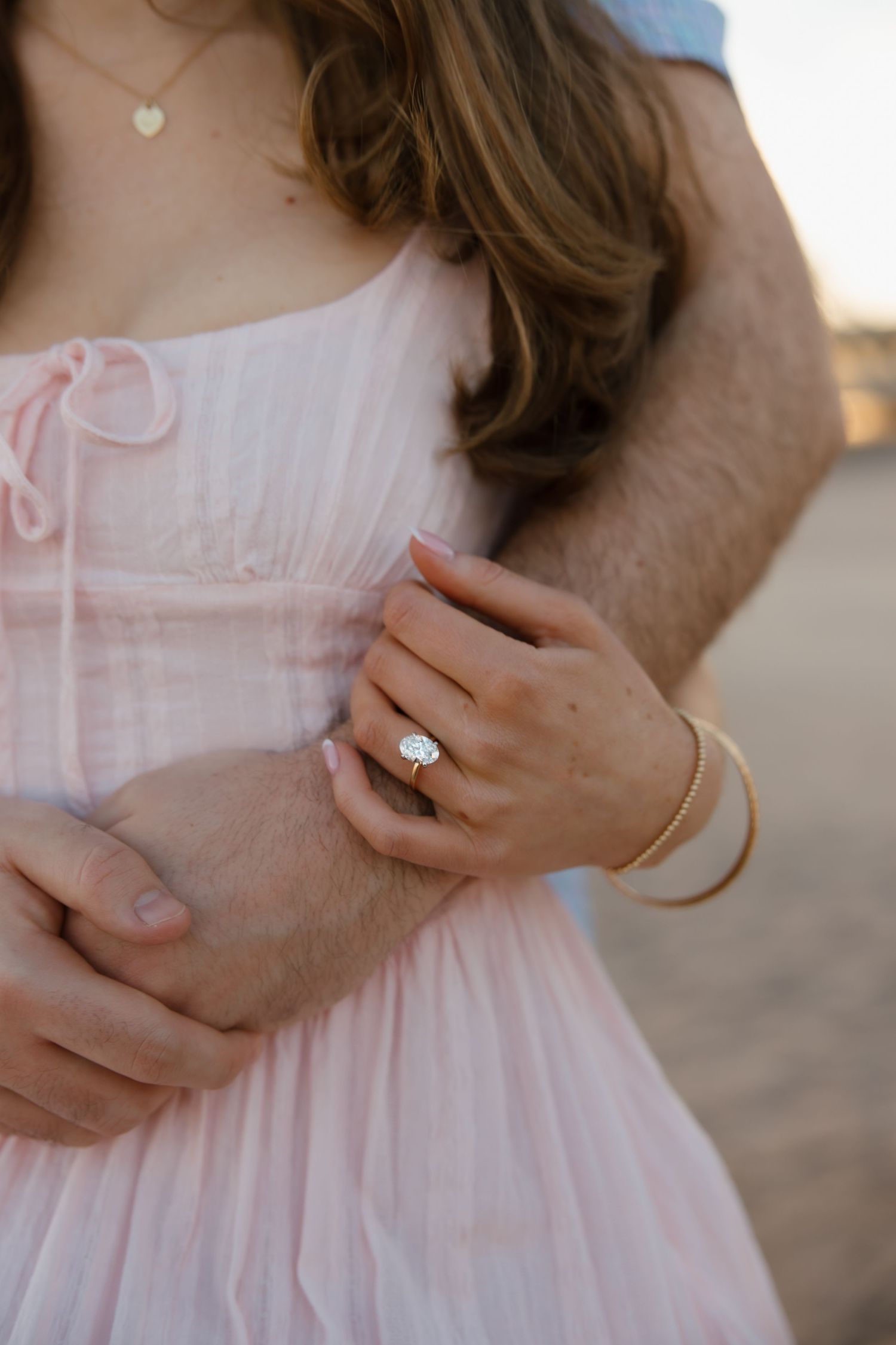 Close-up of a woman's hand with an oval diamond engagement ring, holding a man's arm, wearing a pink dress.