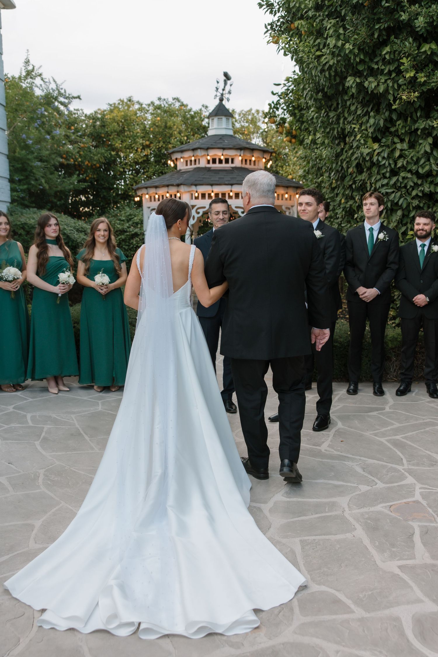 Bride in cathedral veil gown walks toward groom at outdoor ceremony with green bridesmaid dresses.