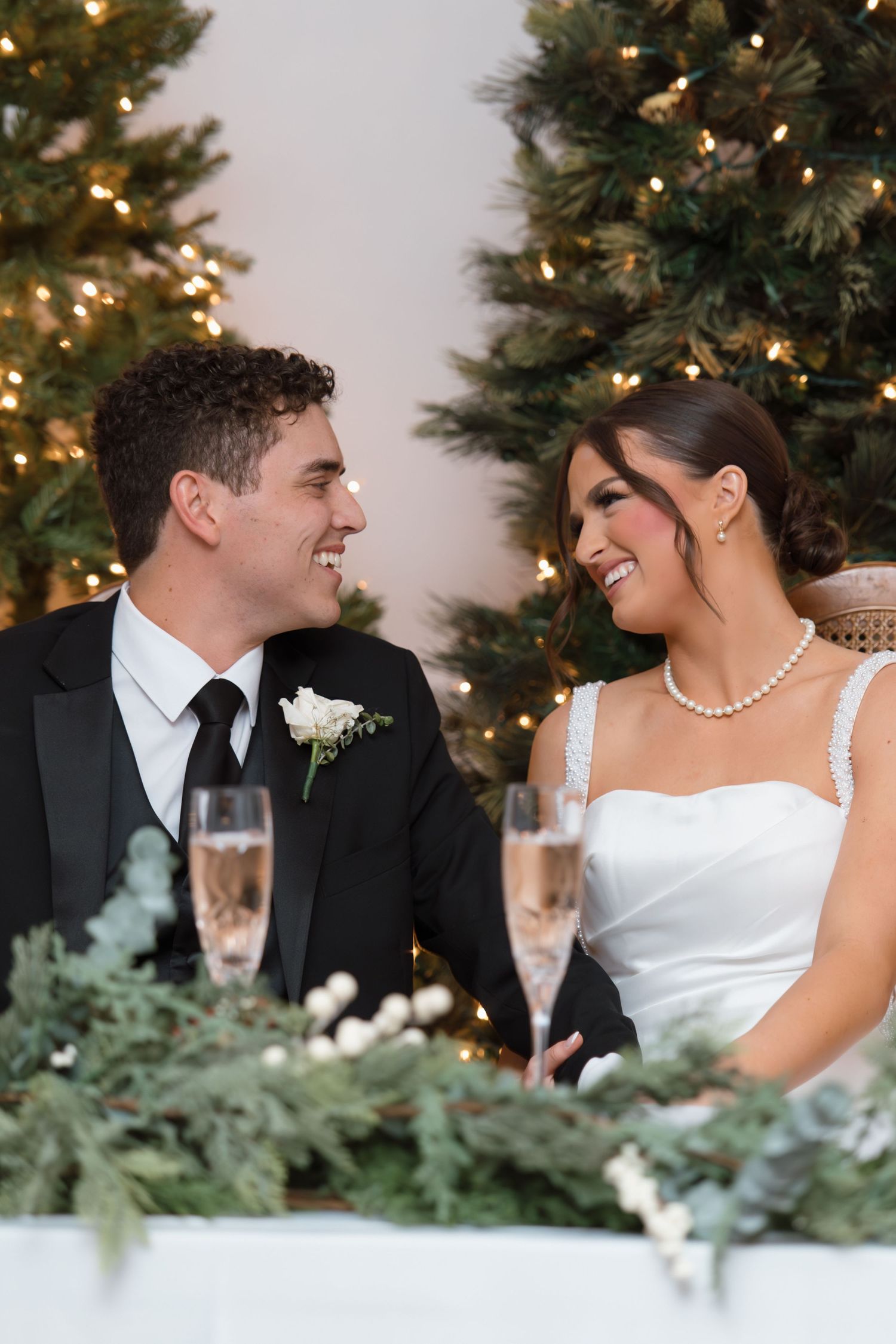 Bride and groom laughing together at sweetheart table with glowing Christmas trees and champagne glasses nearby.