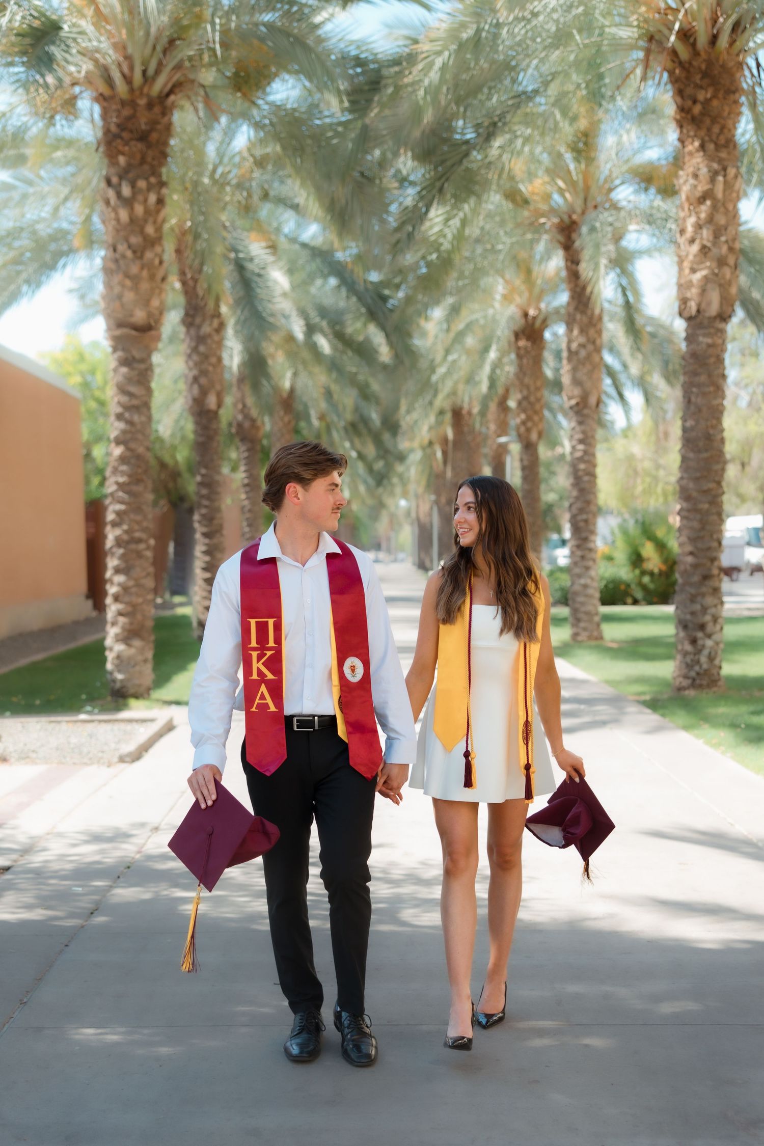 Smiling graduating couple in stoles walking together on sunny palm tree-lined campus path.